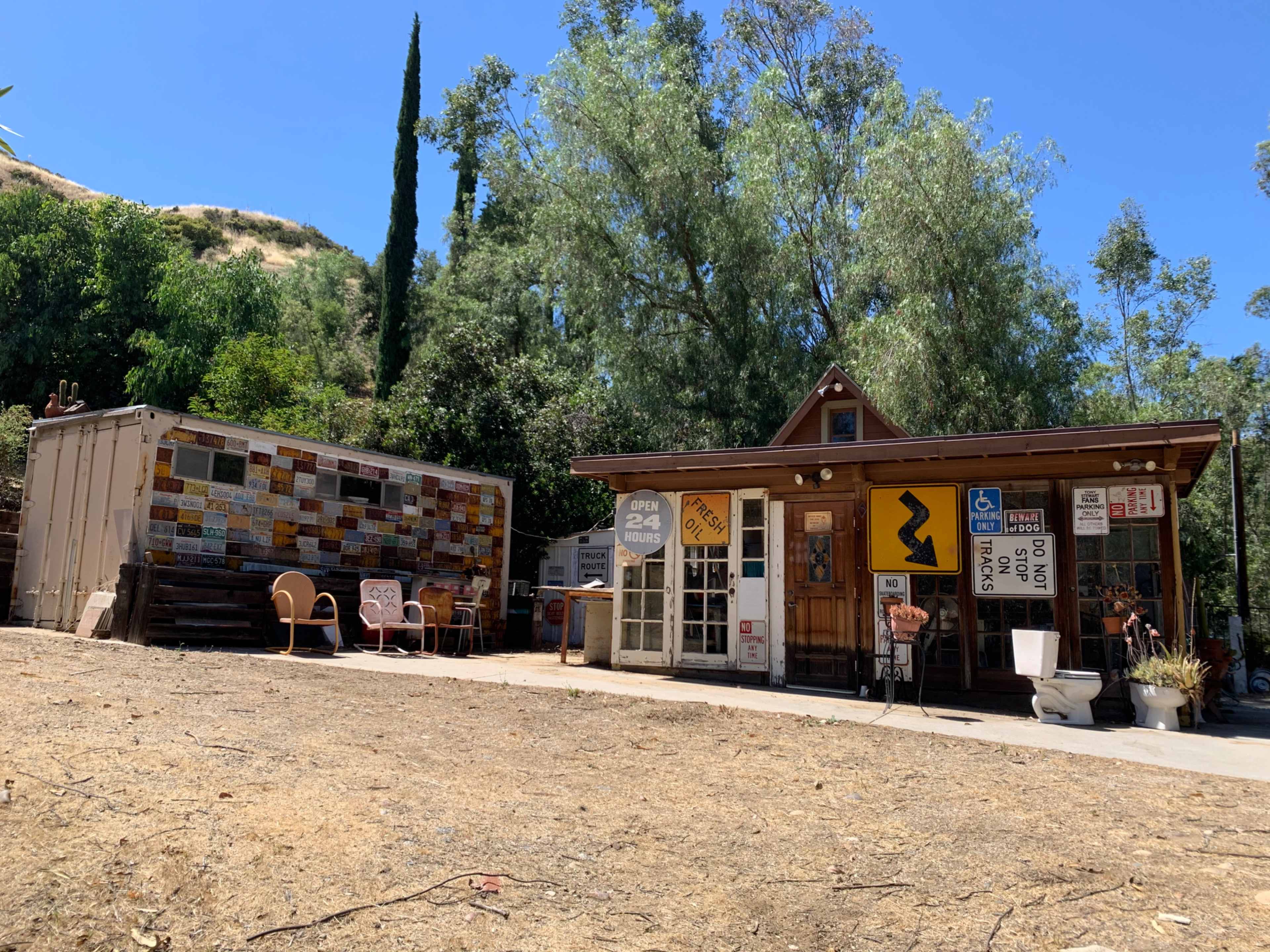 A rustic structure with a wooden facade and various signs, situated next to a shipping container adorned with colorful tiles, surrounded by trees and dry grass.