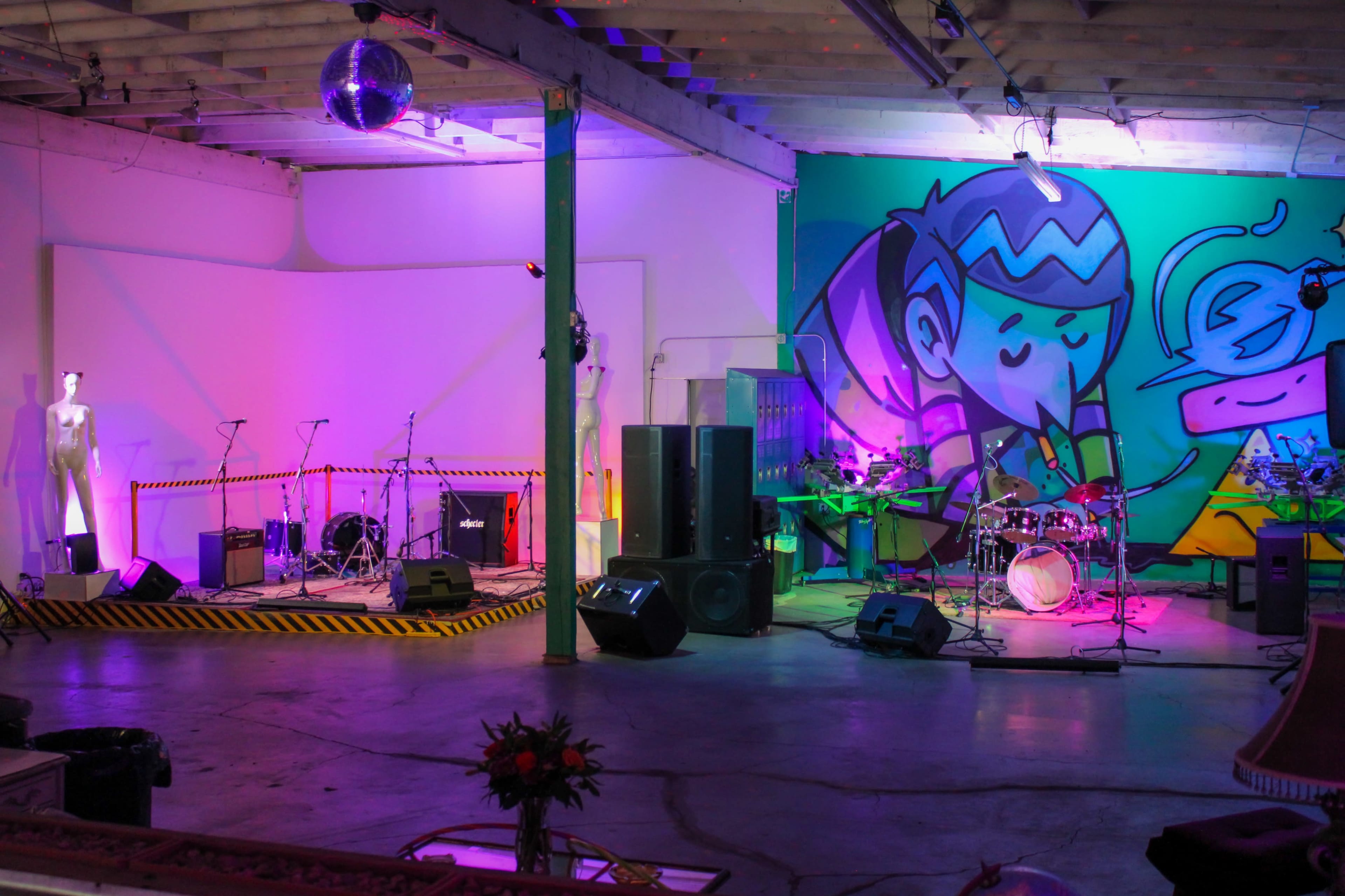 The image shows a colorful indoor venue set up for a performance, featuring a stage with musical instruments, amplifiers, and a large graffiti mural on the wall.