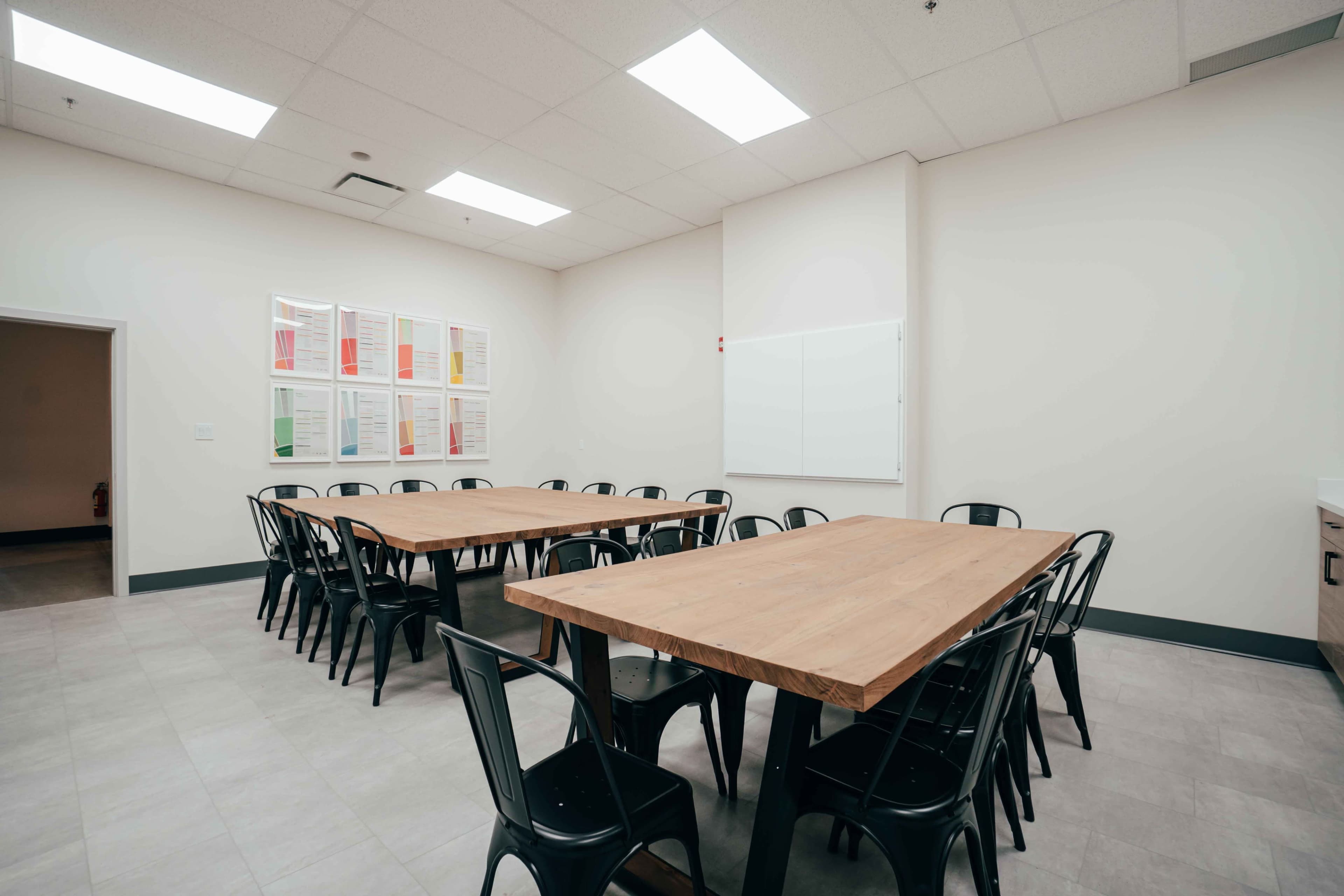 A meeting room features two long wooden tables surrounded by black metal chairs, with colorful posters on the wall and a whiteboard at the front.