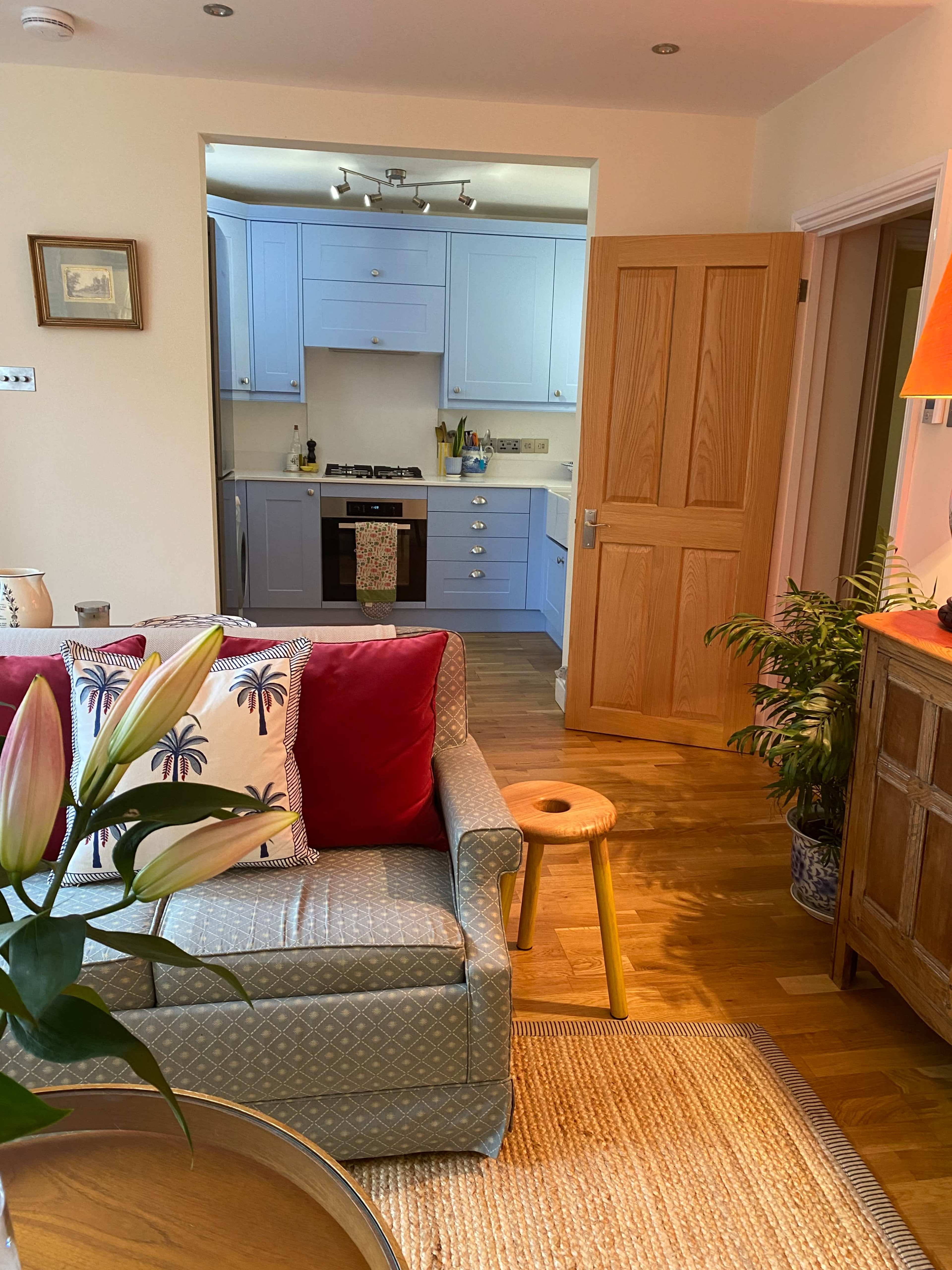 A cozy living room with a light blue kitchen visible in the background and a wooden door leading to a hallway.