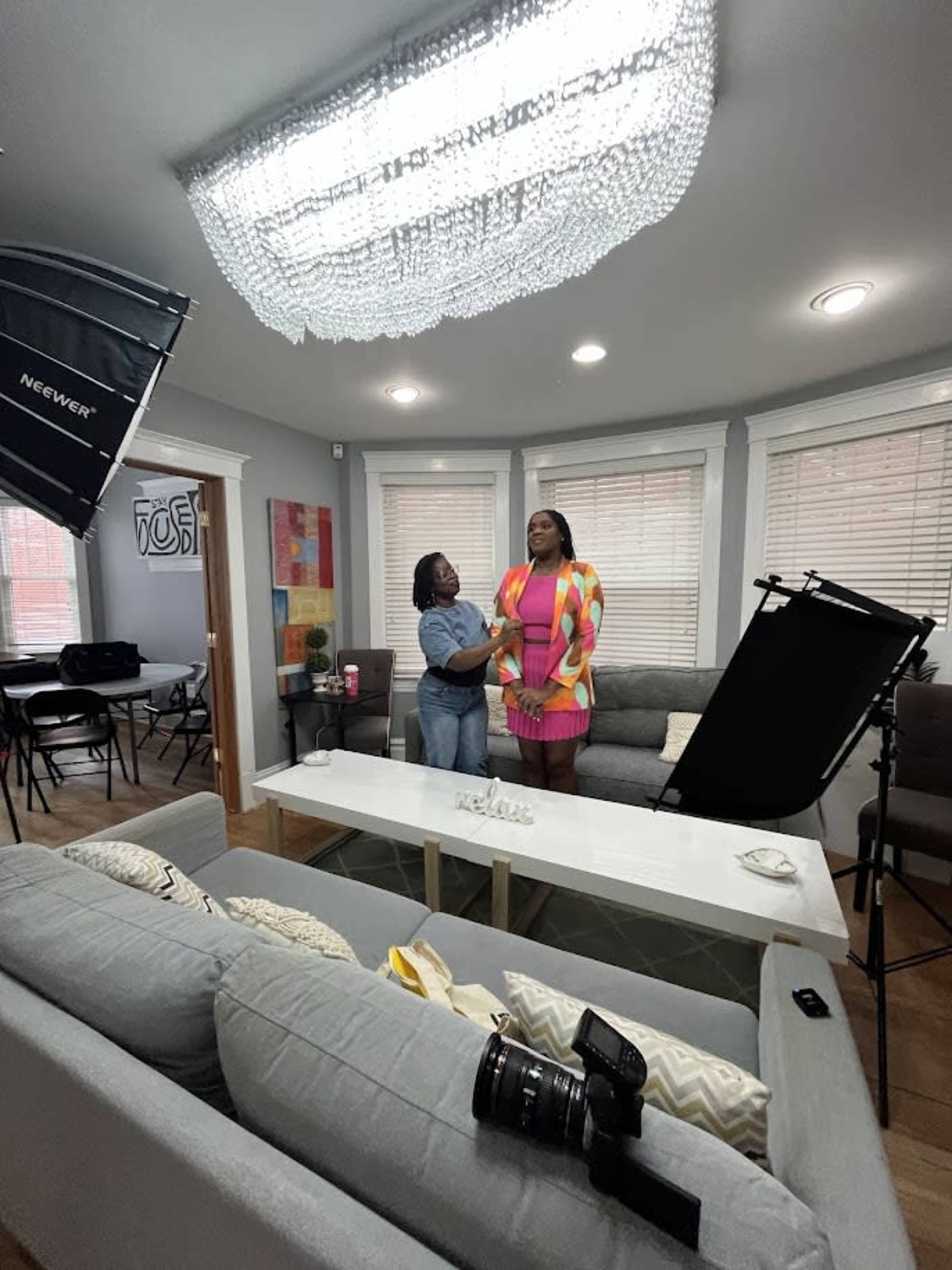 A woman is being styled by another woman in a well-lit living room with a modern decor, featuring a large chandelier and a camera set up nearby.