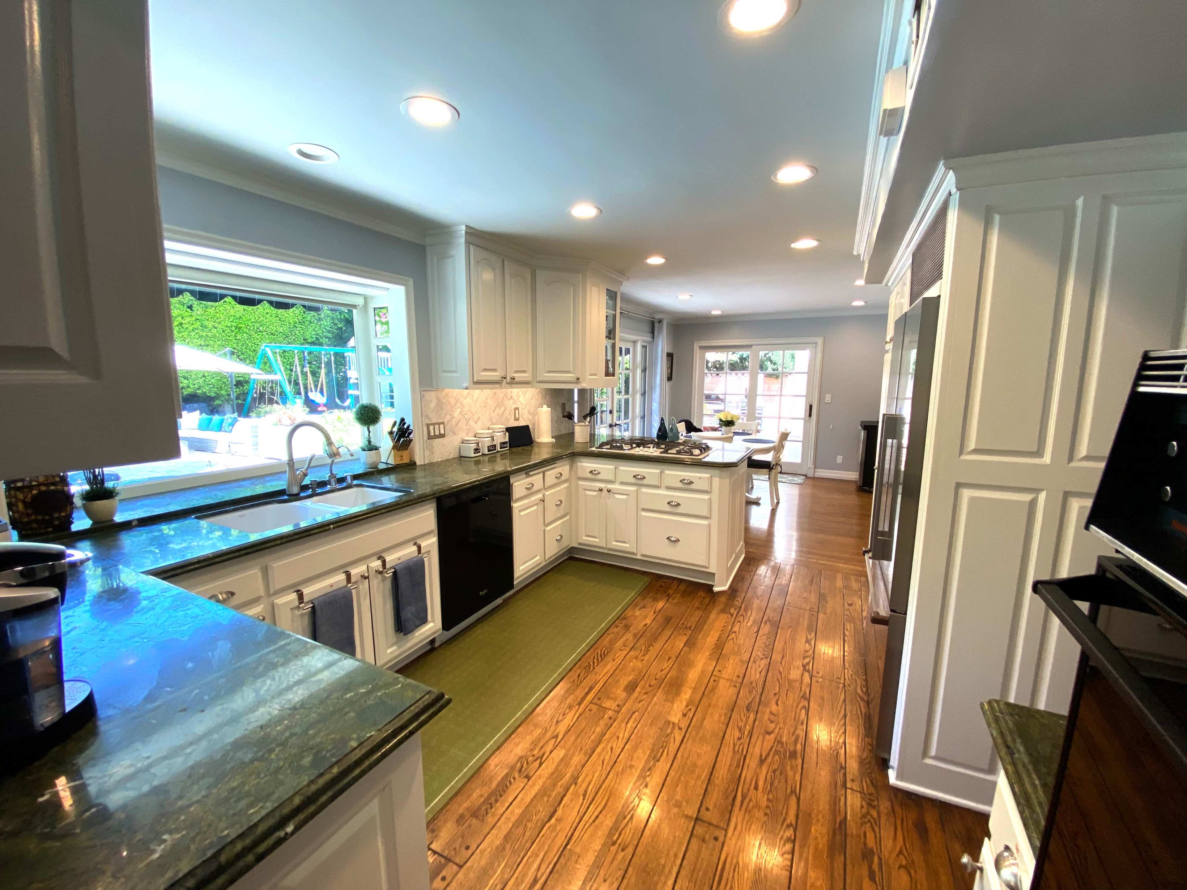 A modern kitchen features white cabinetry, dark countertops, and wooden flooring, with a view of an outdoor area through a large window.