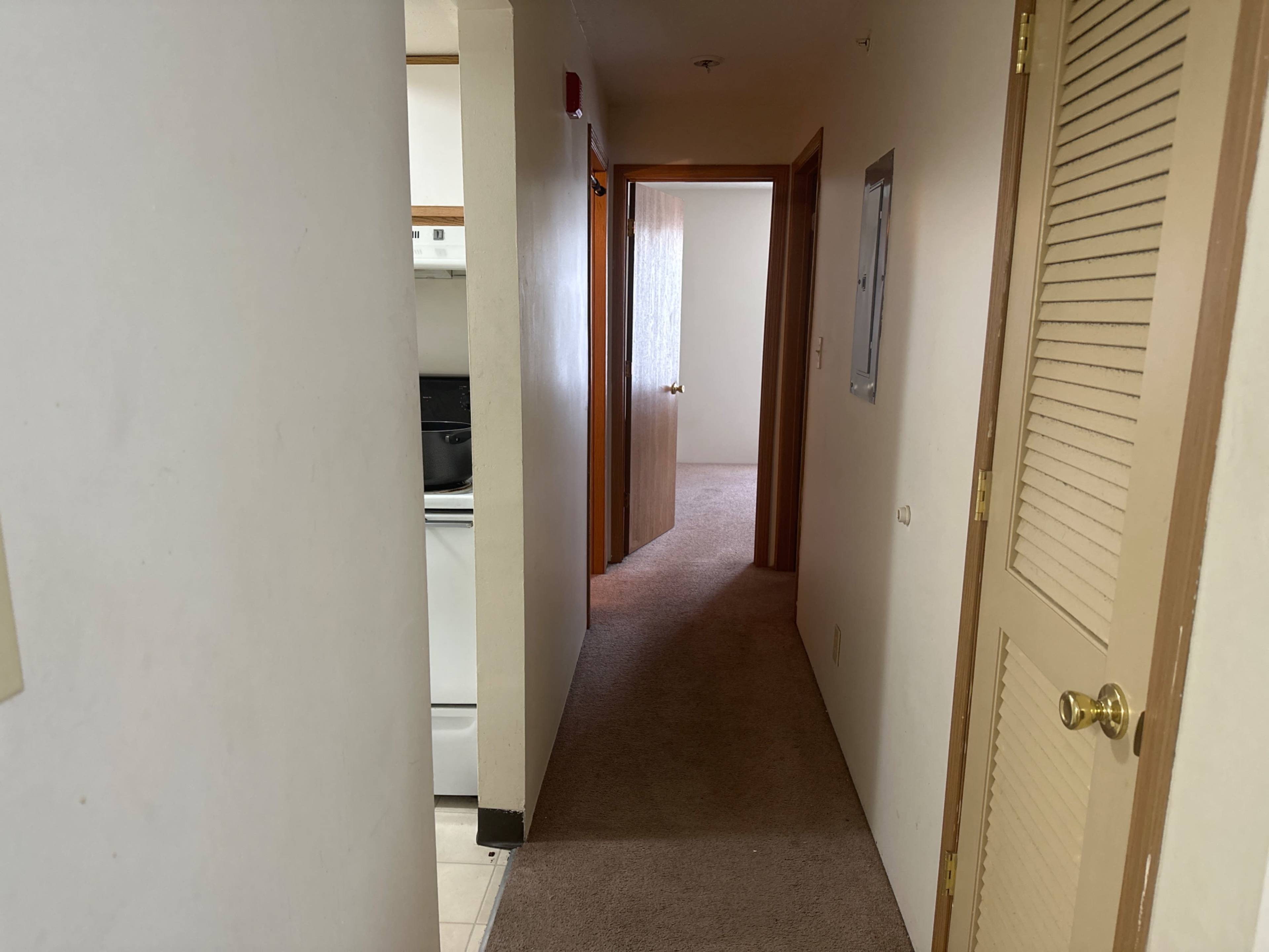 The image shows a narrow hallway leading to two doorways, one opening to a kitchen and the other to a carpeted room.