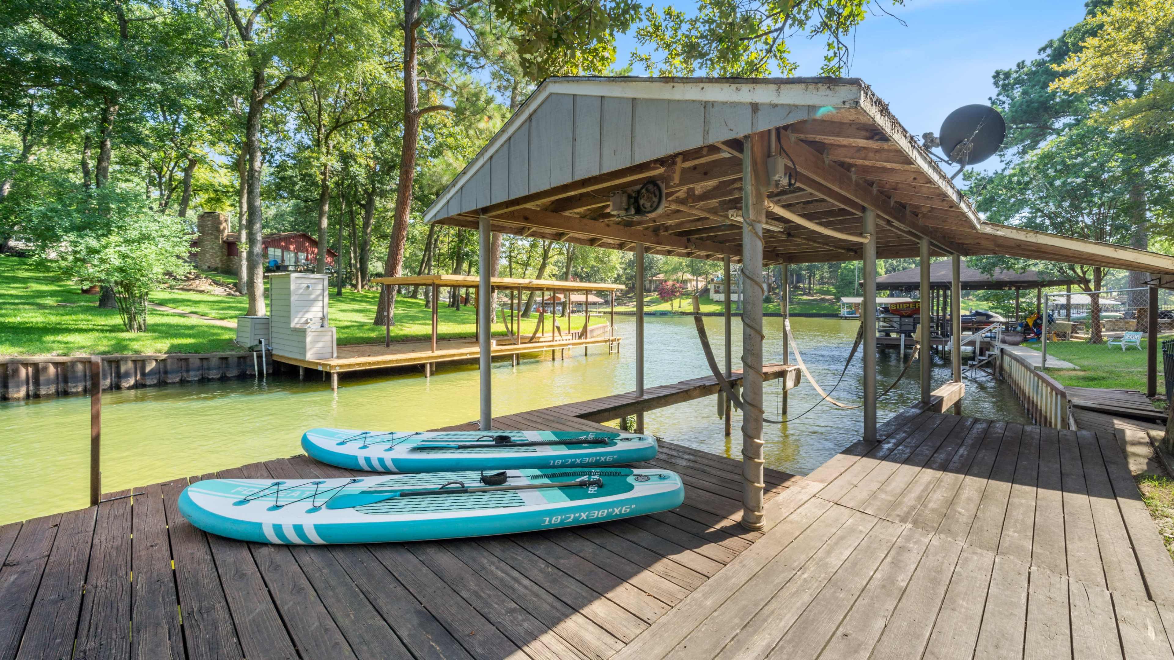 A wooden dock with two stand-up paddleboards rests by a lush green waterway surrounded by trees and nearby docks.