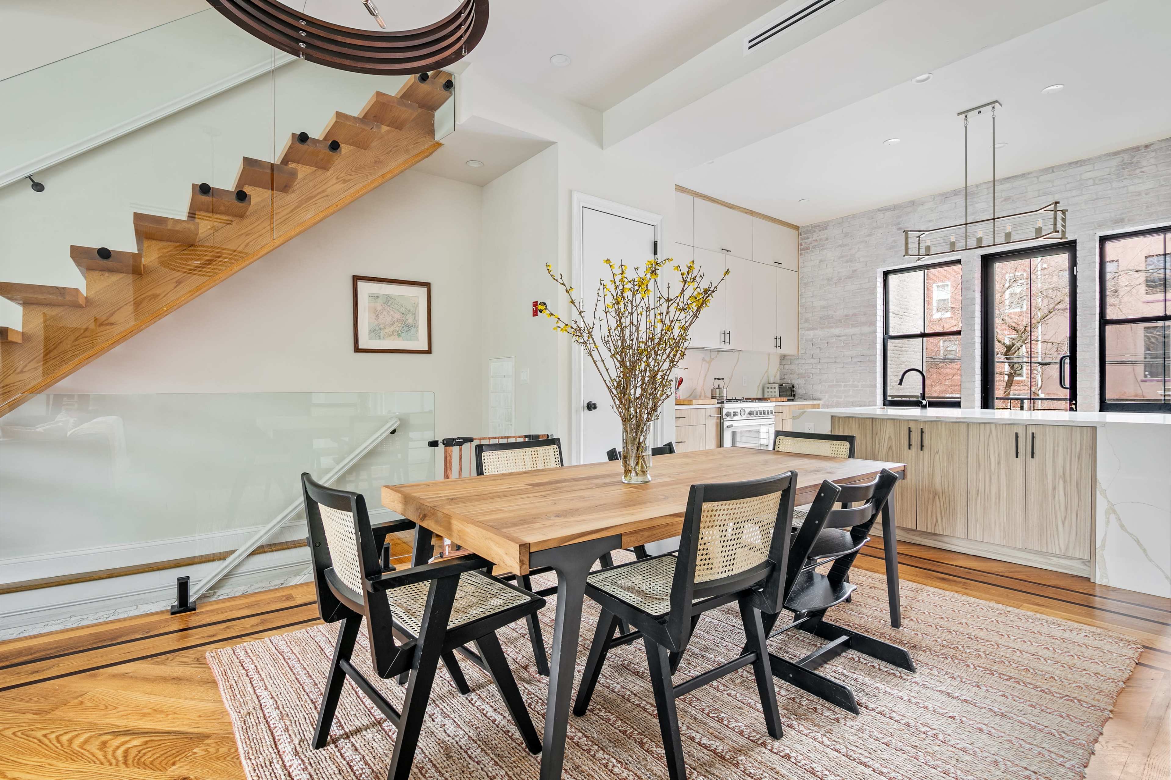 A modern kitchen and dining area features a wooden table surrounded by black chairs, with a staircase in the background and a vase of flowers on the table.