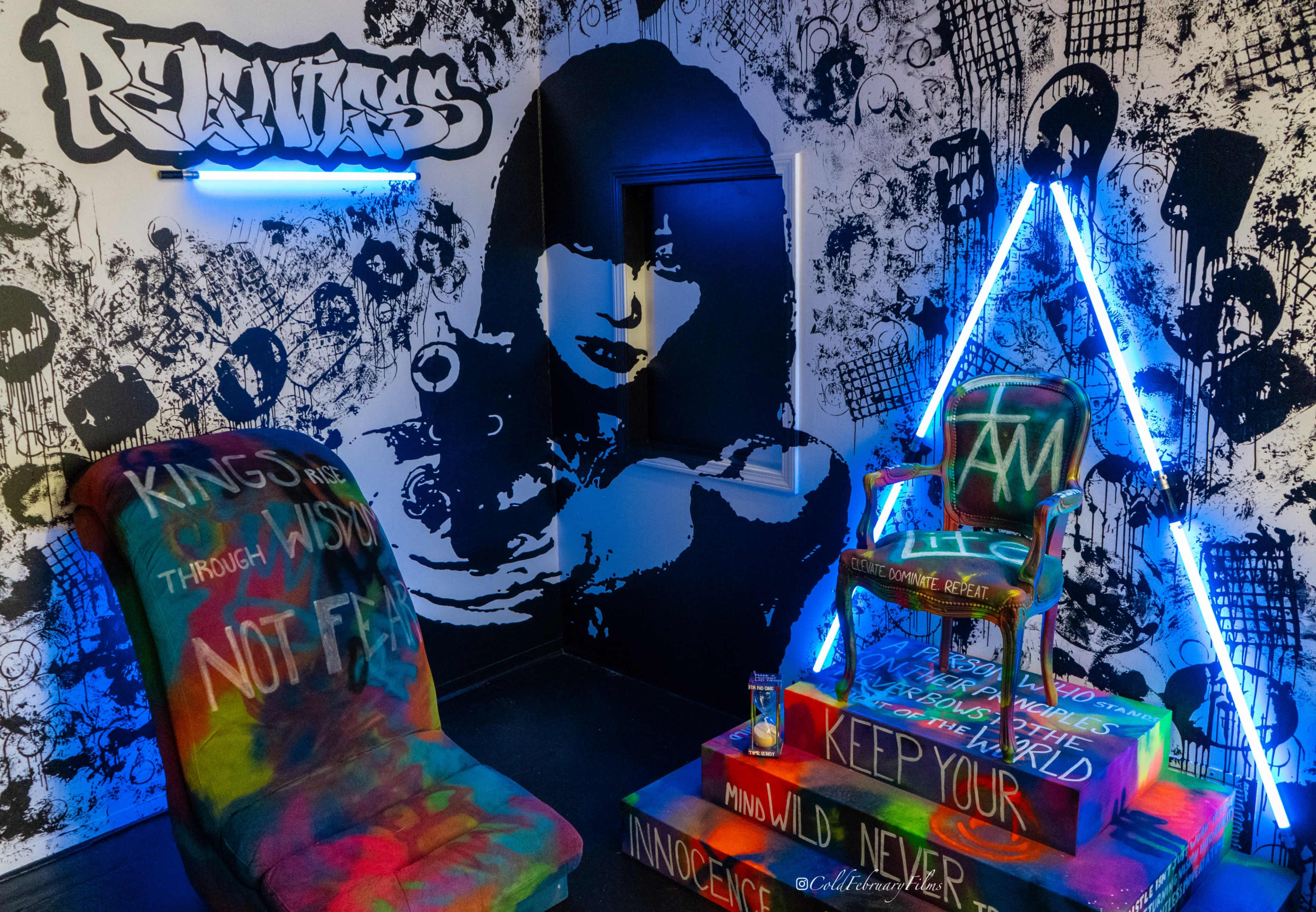 The image shows a creatively designed room with graffiti-covered walls and colorful furniture, featuring a neon light and an artistic portrait.