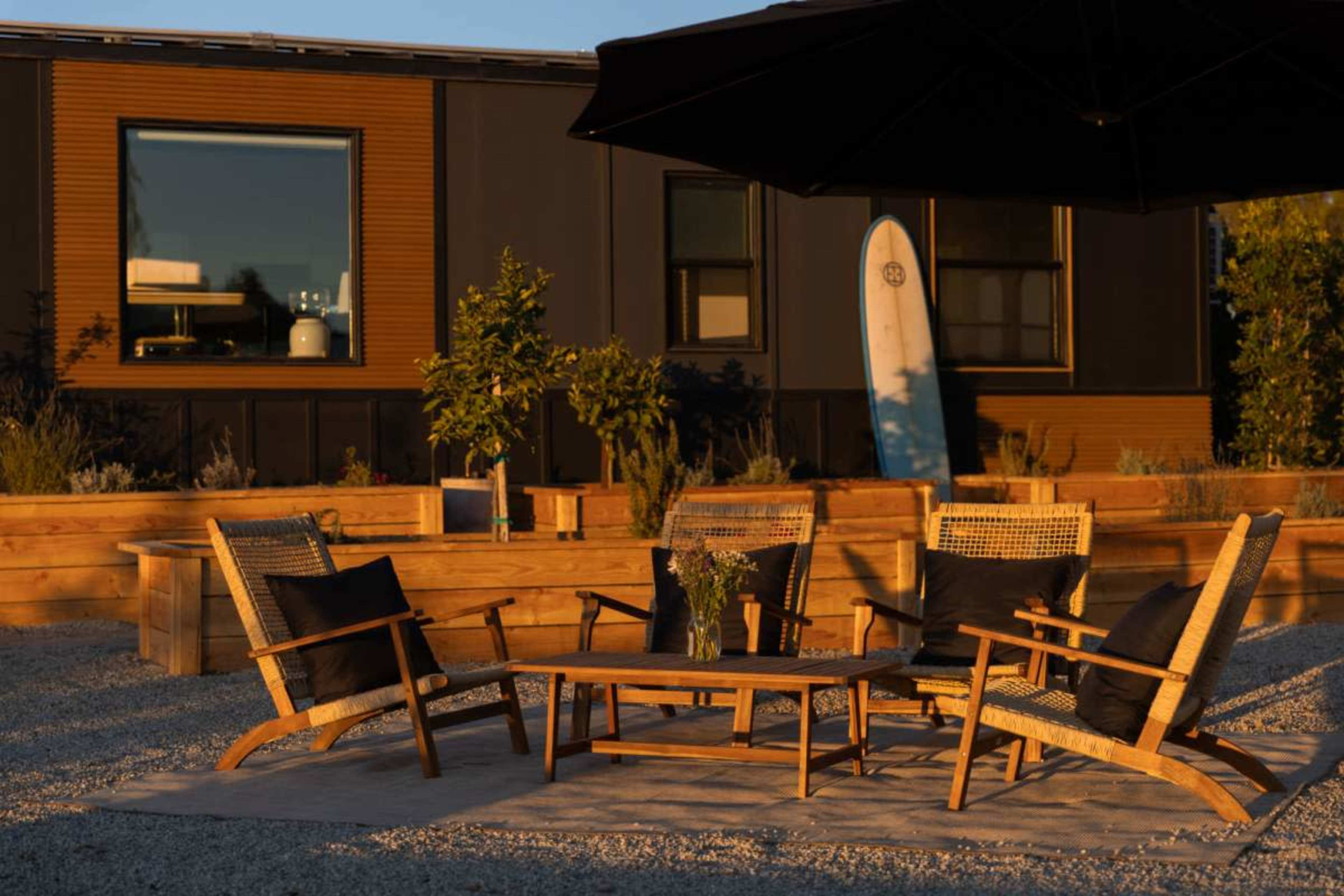 The scene depicts a cozy outdoor seating area with wooden furniture, a large umbrella, and a surfboard leaning against a building in the background.