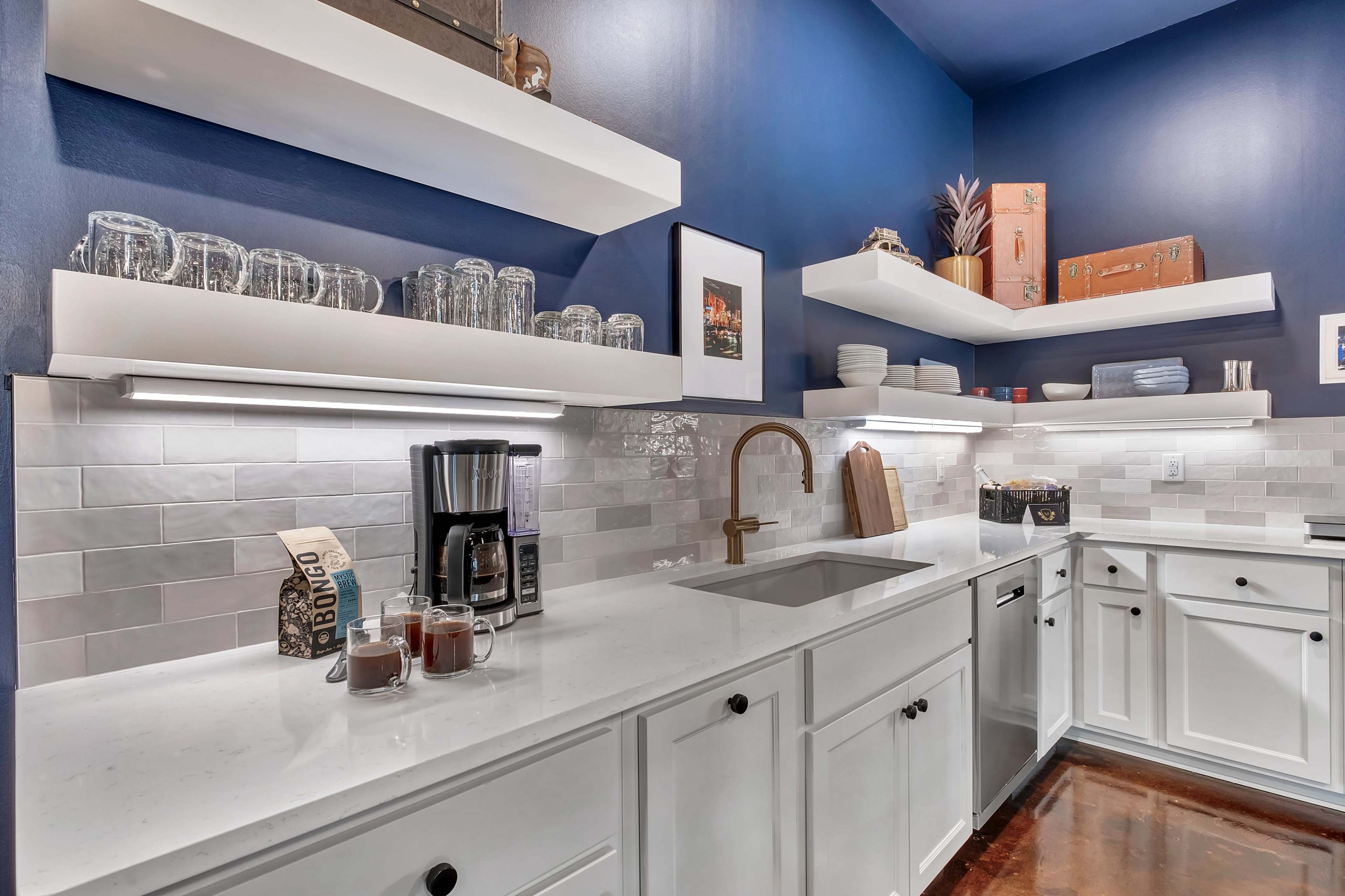 The image shows a modern kitchen with open shelving displaying glassware and dishware, a sleek sink, a coffee maker, and a dark blue wall.