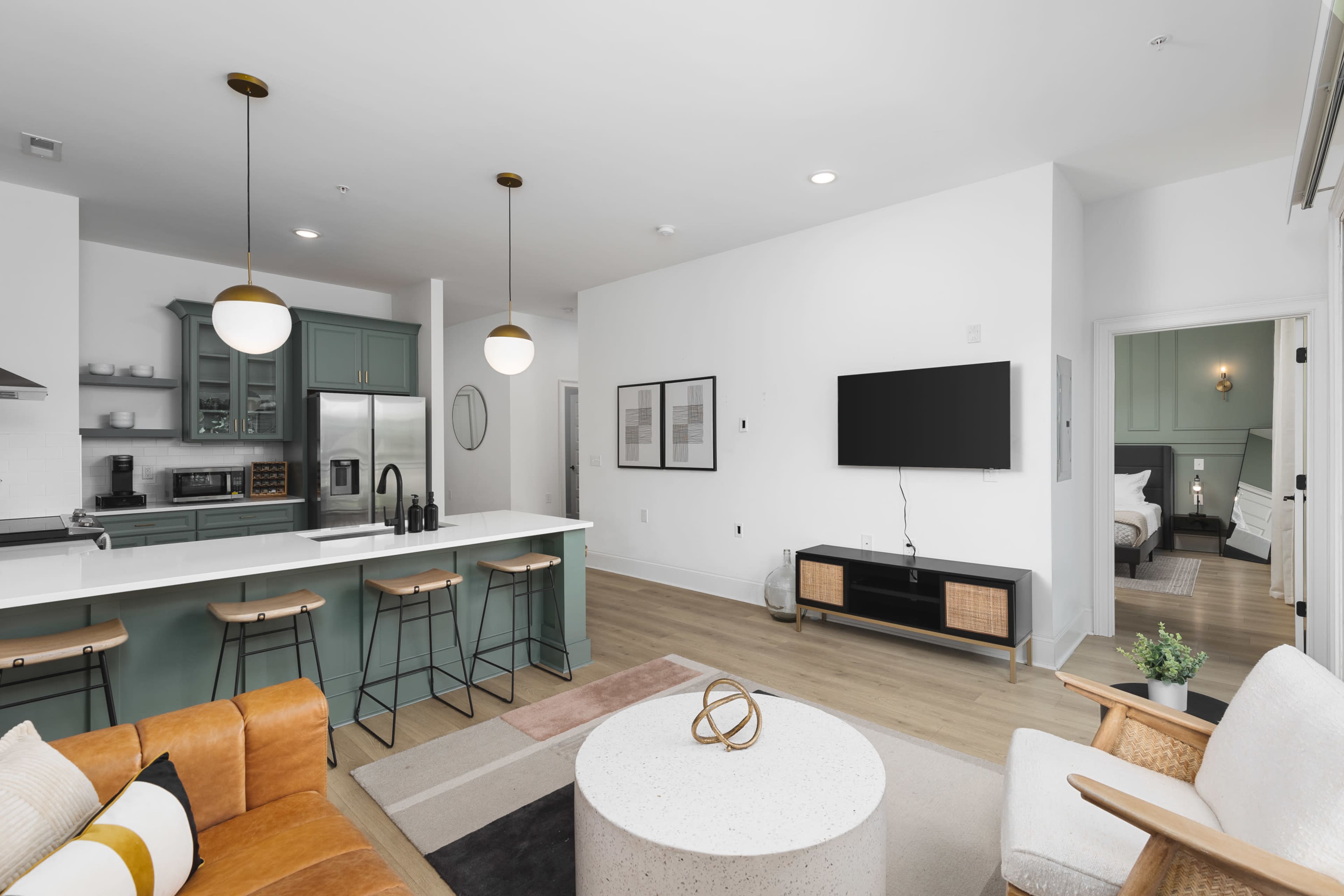 A modern living room features a connected kitchen area with green cabinetry, a large island with bar stools, and a flat-screen TV mounted on a wall opposite a cozy seating arrangement.