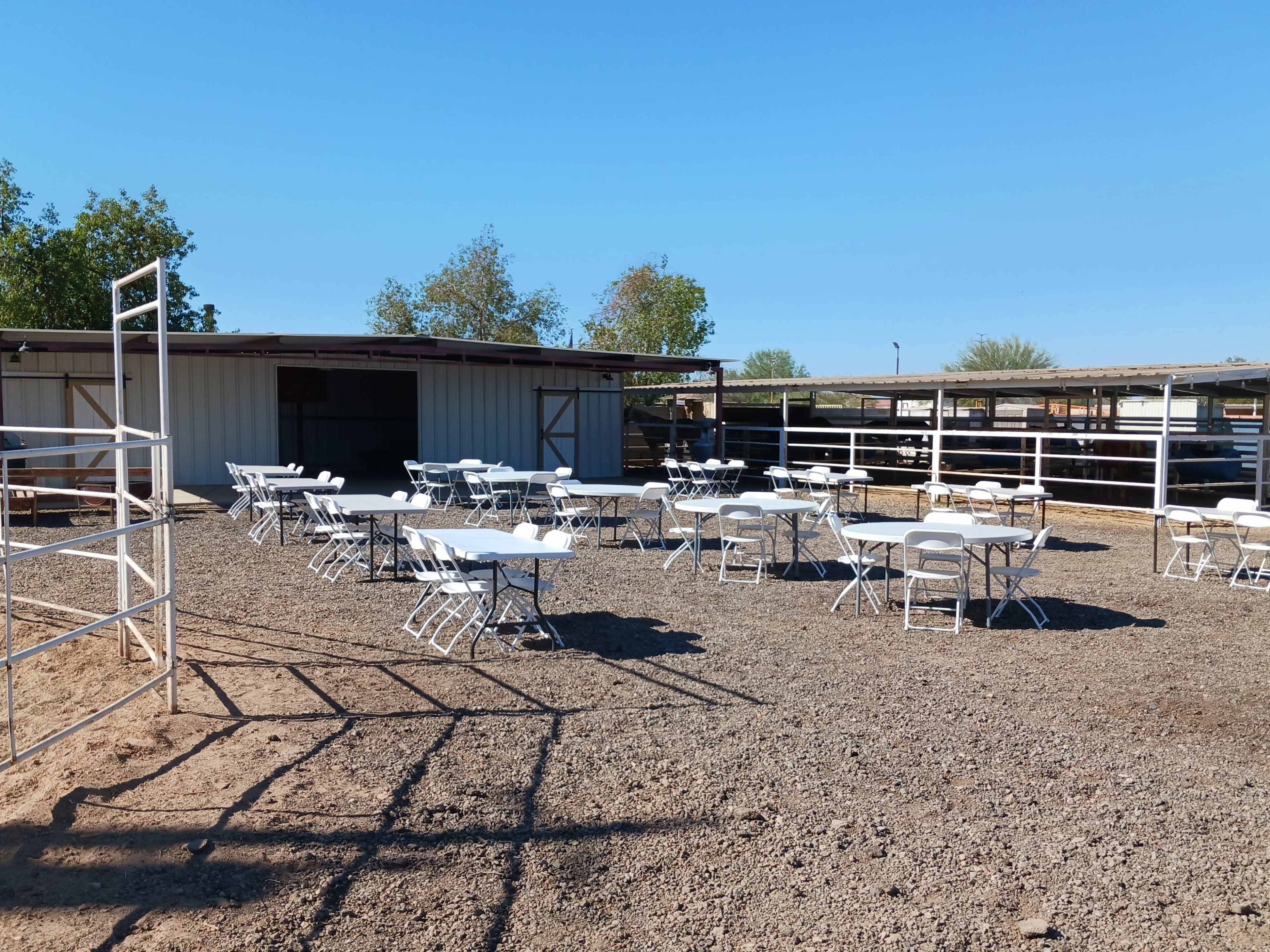 A gravel area with white folding tables set up in front of a metal barn and a fenced enclosure.