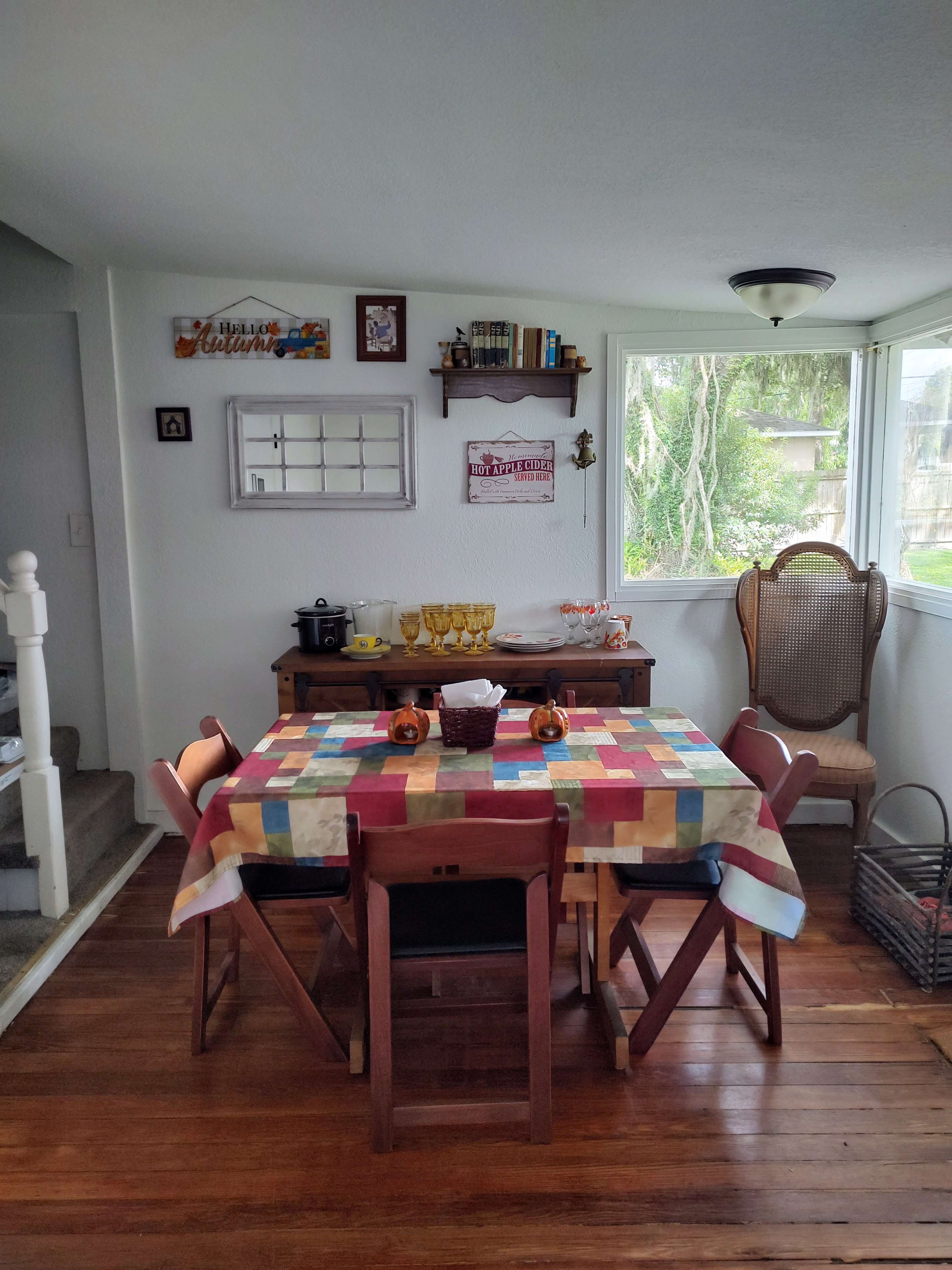 A dining area with a colorful tablecloth-covered table surrounded by wooden chairs, a sideboard with various dishes, and large windows letting in natural light.