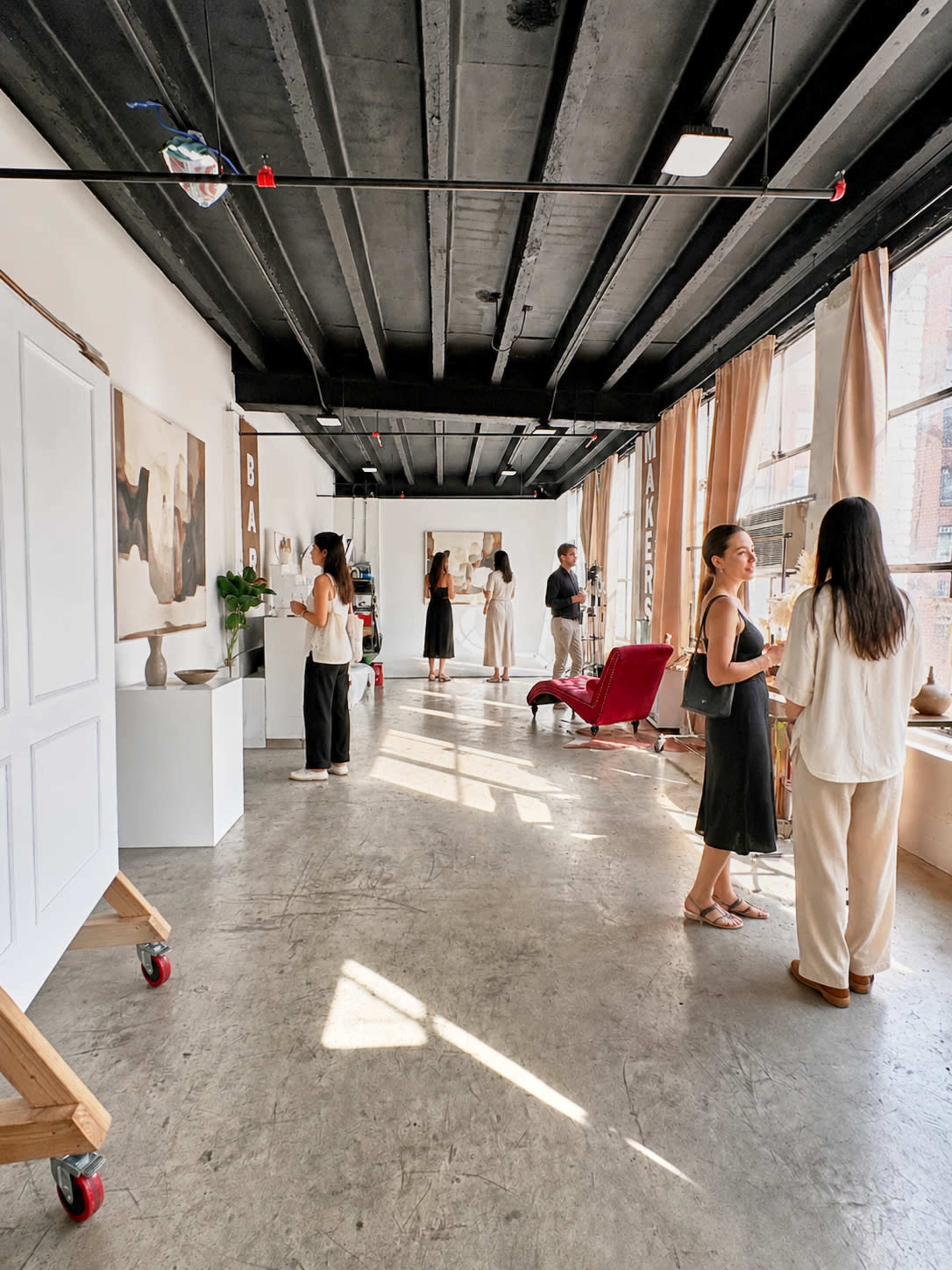 The image shows a spacious, modern studio with several people interacting, showcasing art and decorative items against a backdrop of large windows and exposed beams.