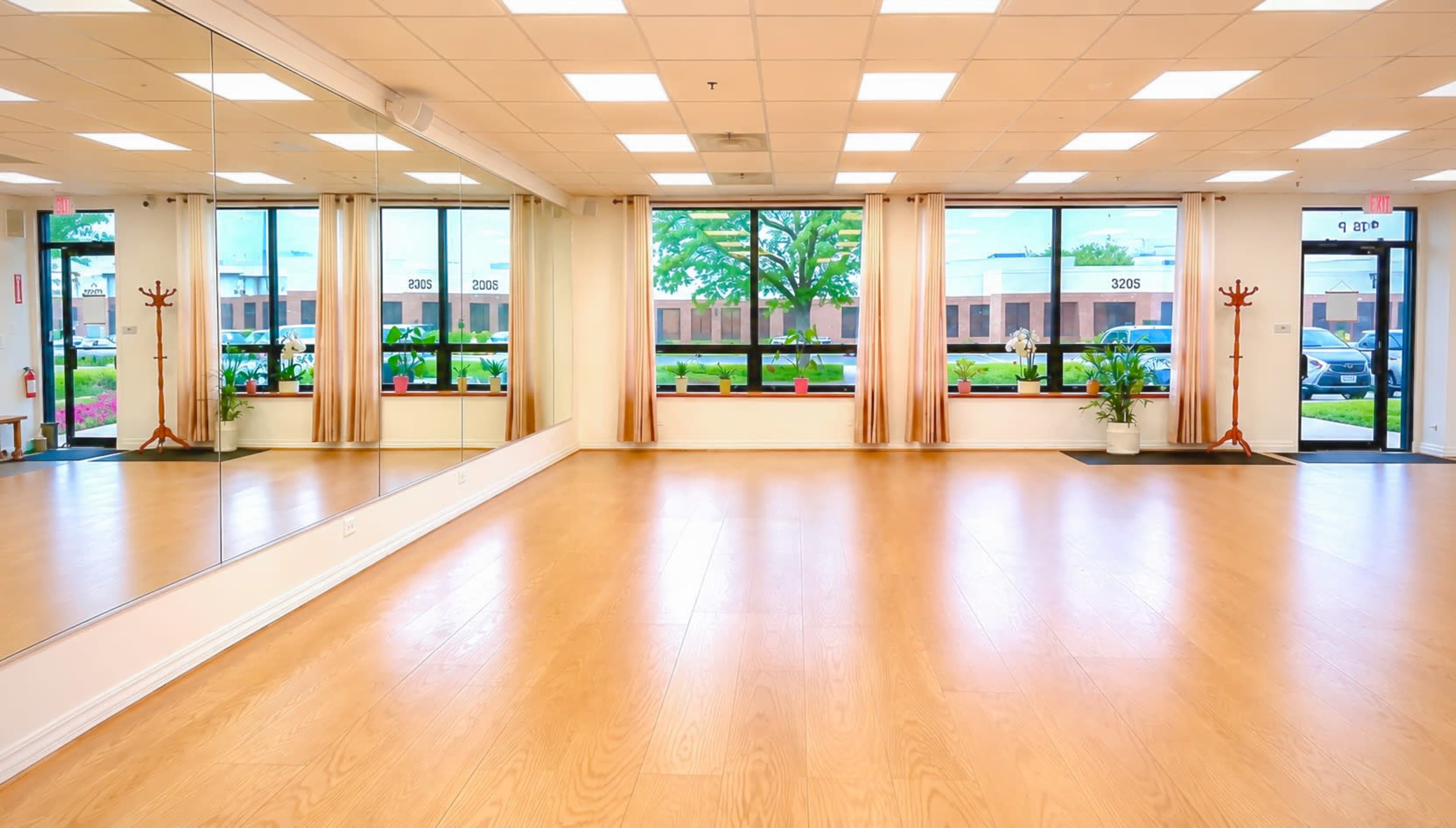 The image shows an interior space with wooden flooring, large windows letting in natural light, and a mirror along one wall.