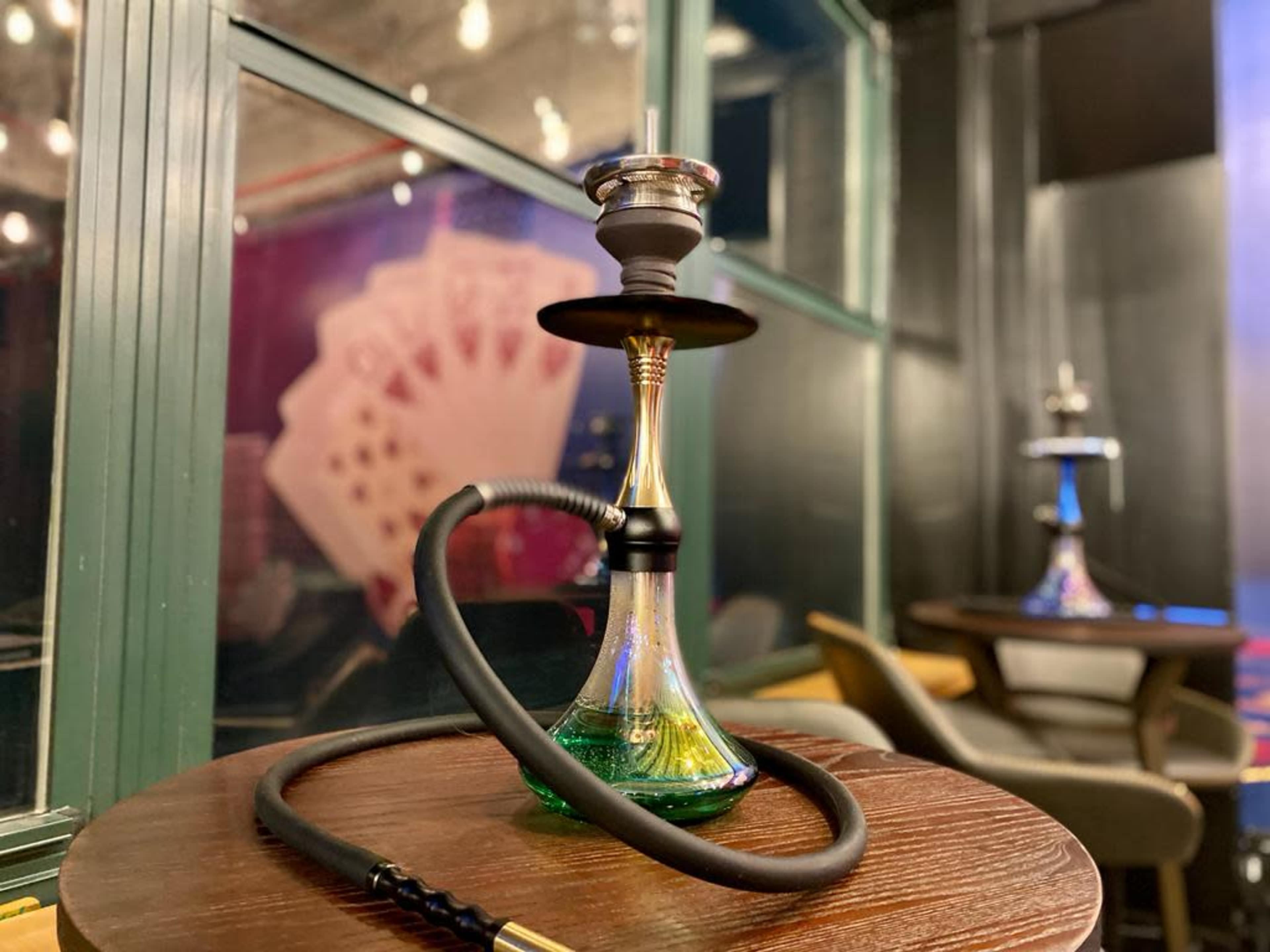 A hookah with a colorful base sits on a wooden table, with playing cards displayed in the background.
