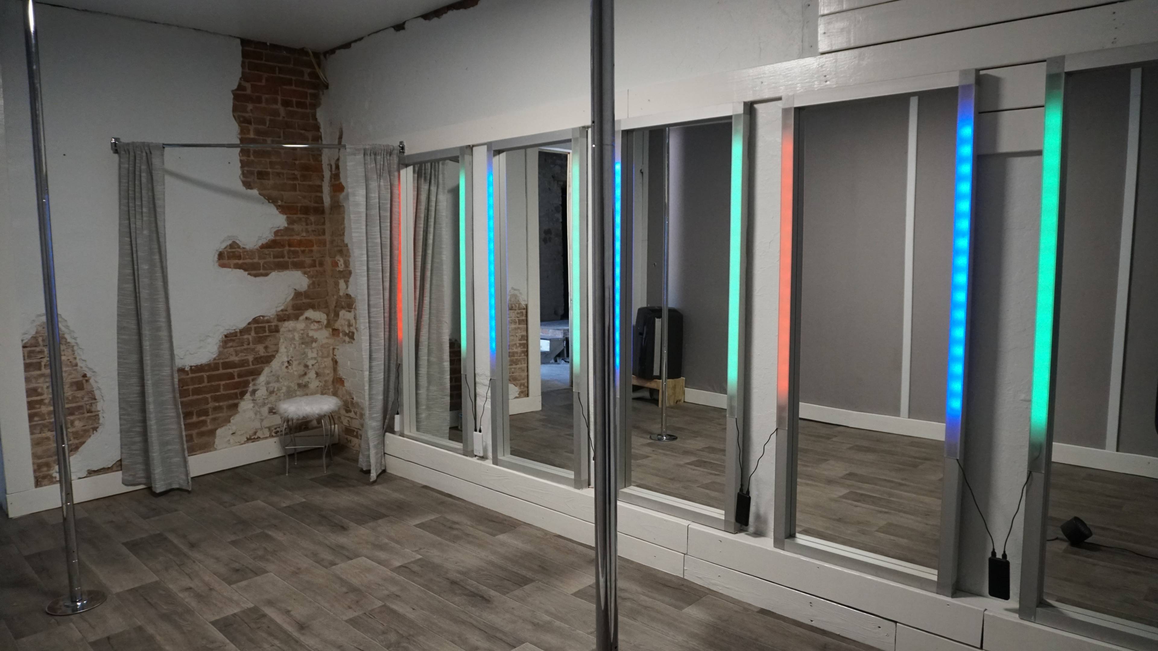 A room with a pole for dancing, illuminated mirrors, and colored LED lights, featuring a rustic wall with exposed bricks.