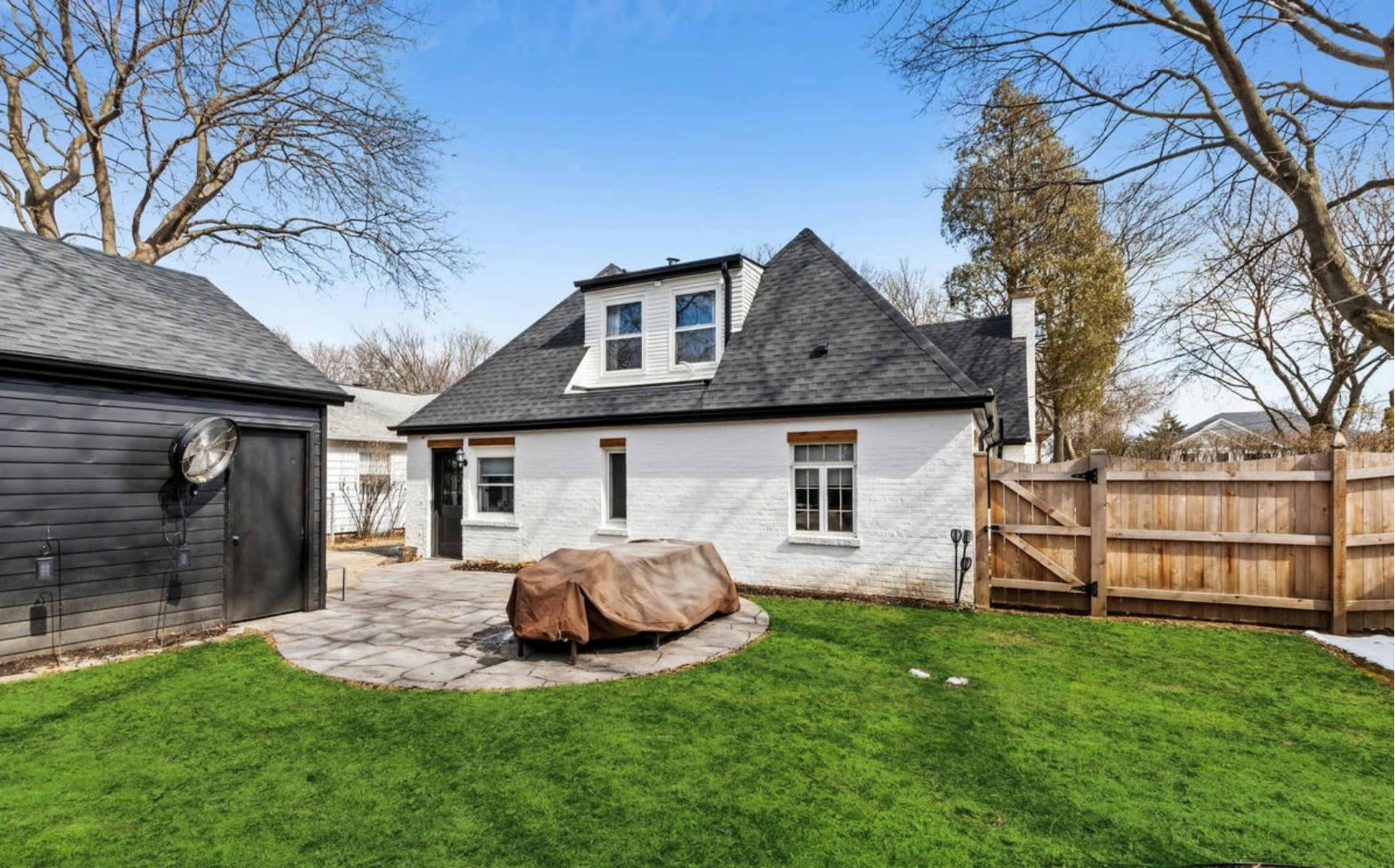 A backyard with a patio featuring a covered outdoor table, surrounded by grass and bordered by a wooden fence, with a house and shed in the background.