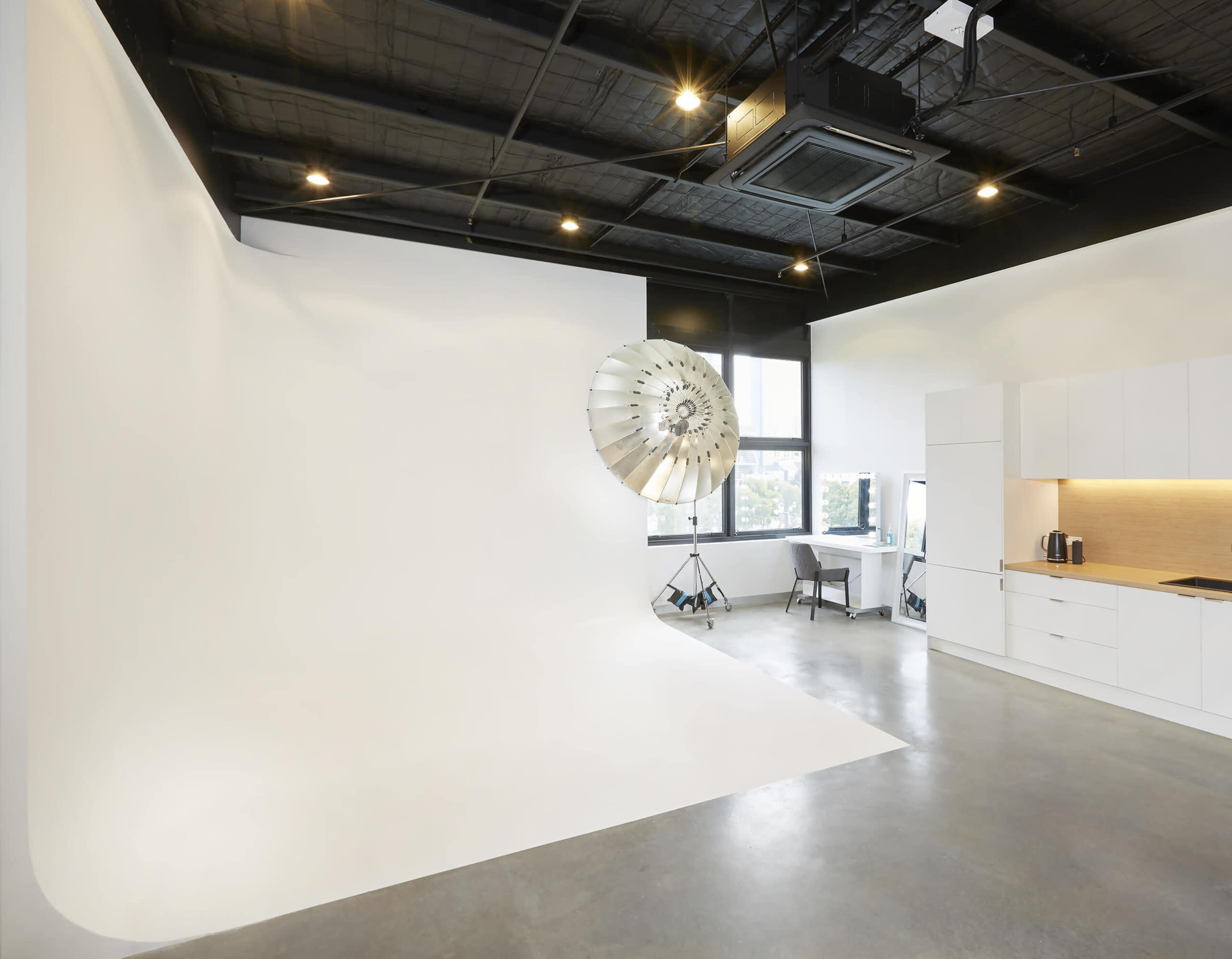 A minimalist photography studio featuring a seamless white backdrop, a large reflector, and a modern kitchen area with cabinetry.