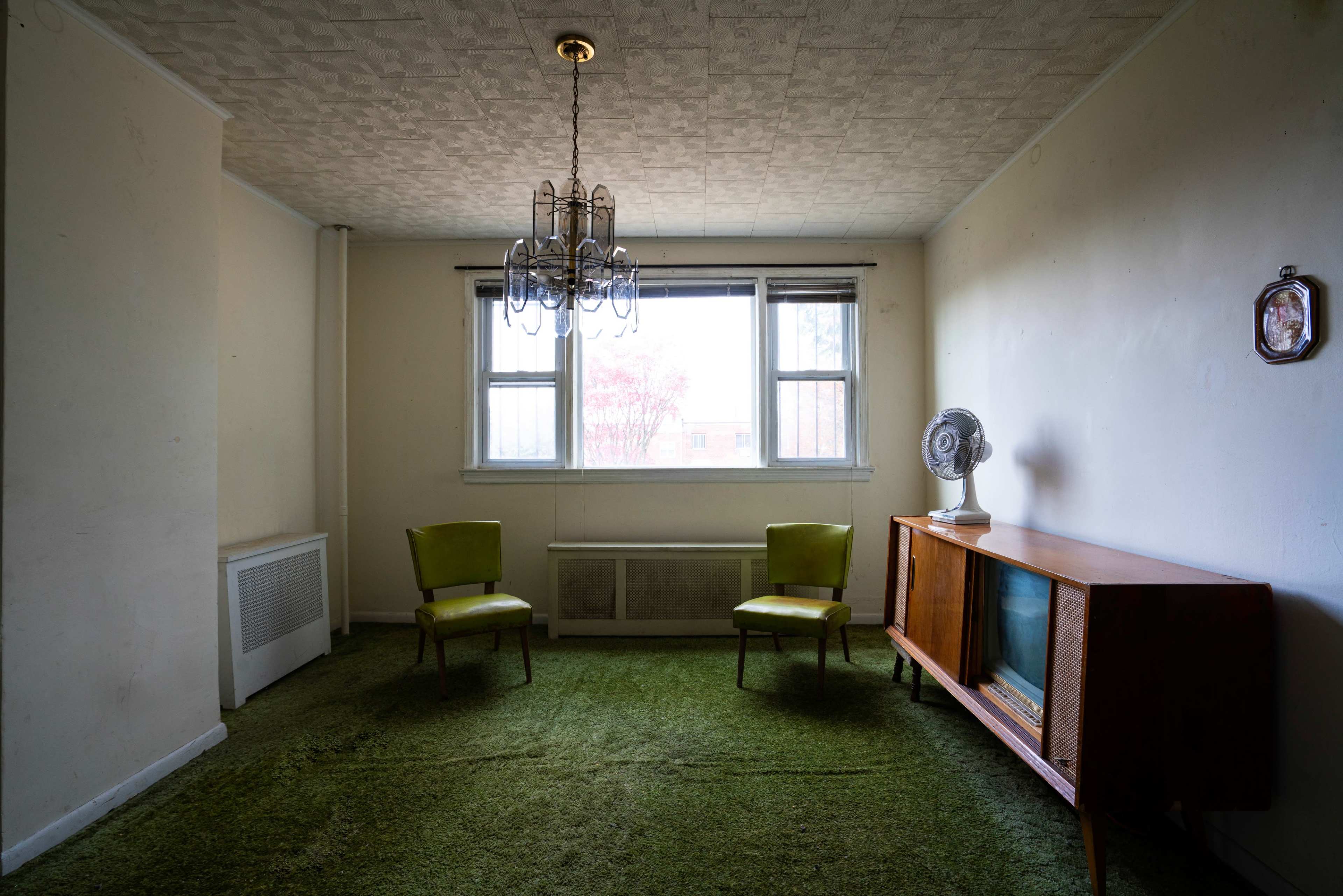 The image shows an empty room with two green chairs, a wooden television cabinet, a ceiling chandelier, and a window overlooking an outdoor view.