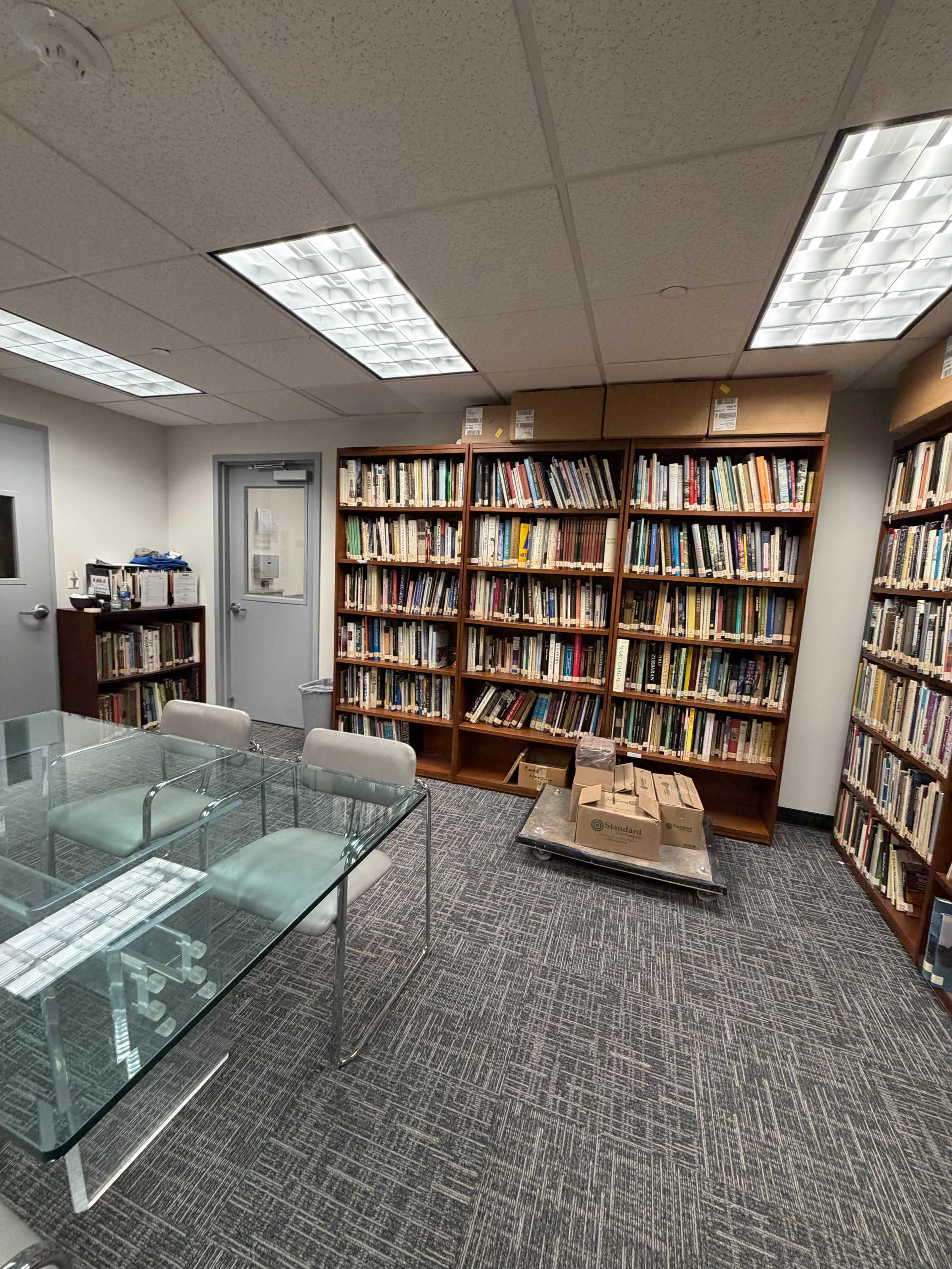 The image shows a small room with bookshelves filled with books on two walls, a glass table with chairs in the center, and a stack of boxes on the floor.