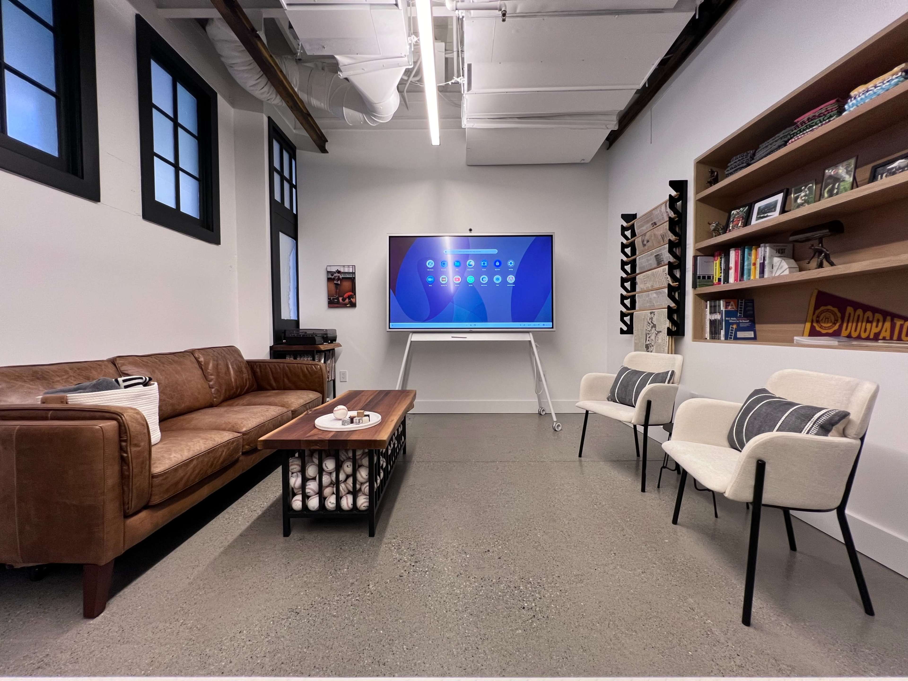 The image shows a modern office space featuring a brown leather sofa, two white chairs, a coffee table, a wall-mounted display screen, and a bookshelf against a white wall.