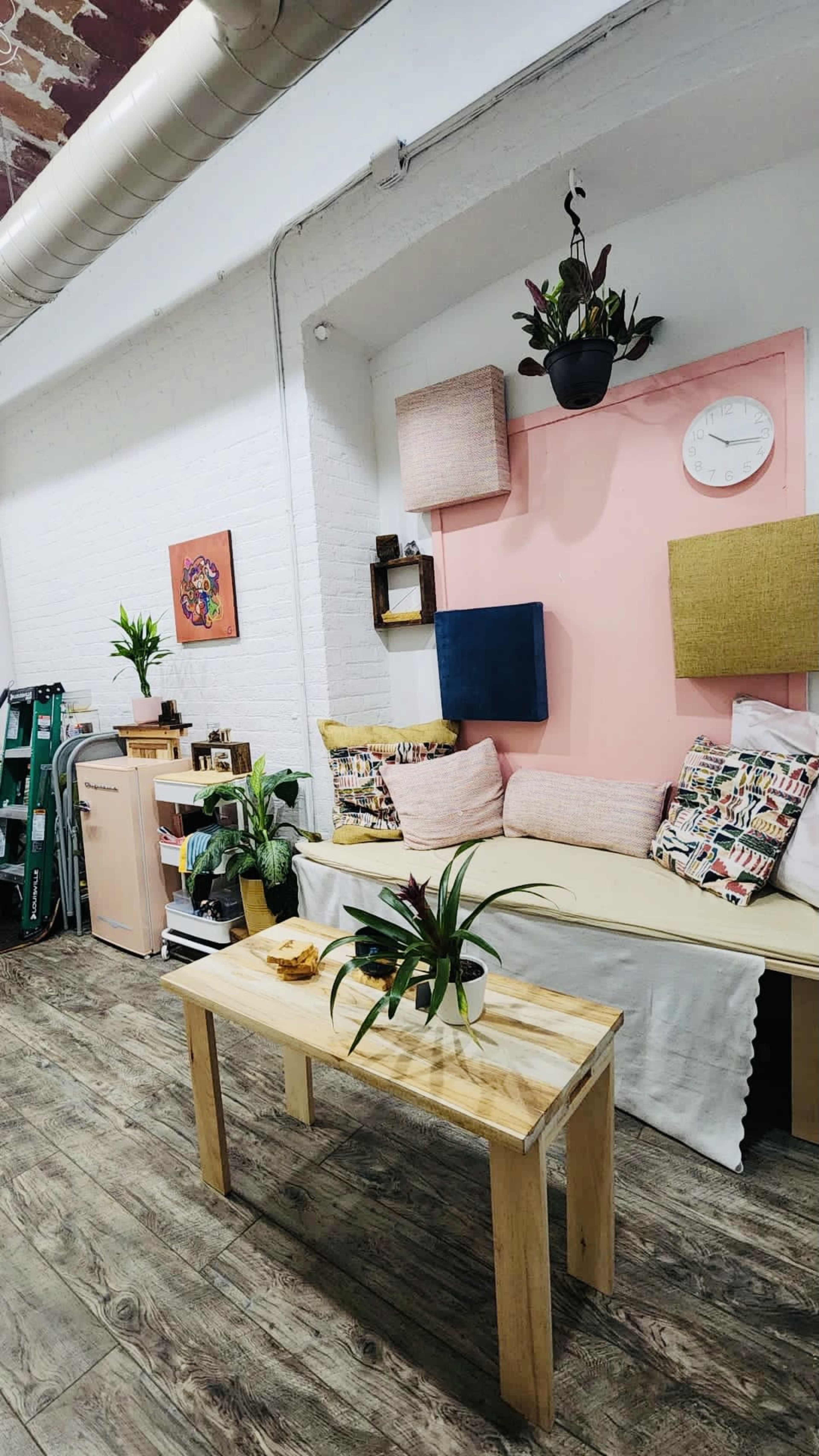 A cozy living space featuring a neutral sofa adorned with decorative cushions, a wooden coffee table, and various plants against a backdrop of white and pink walls.