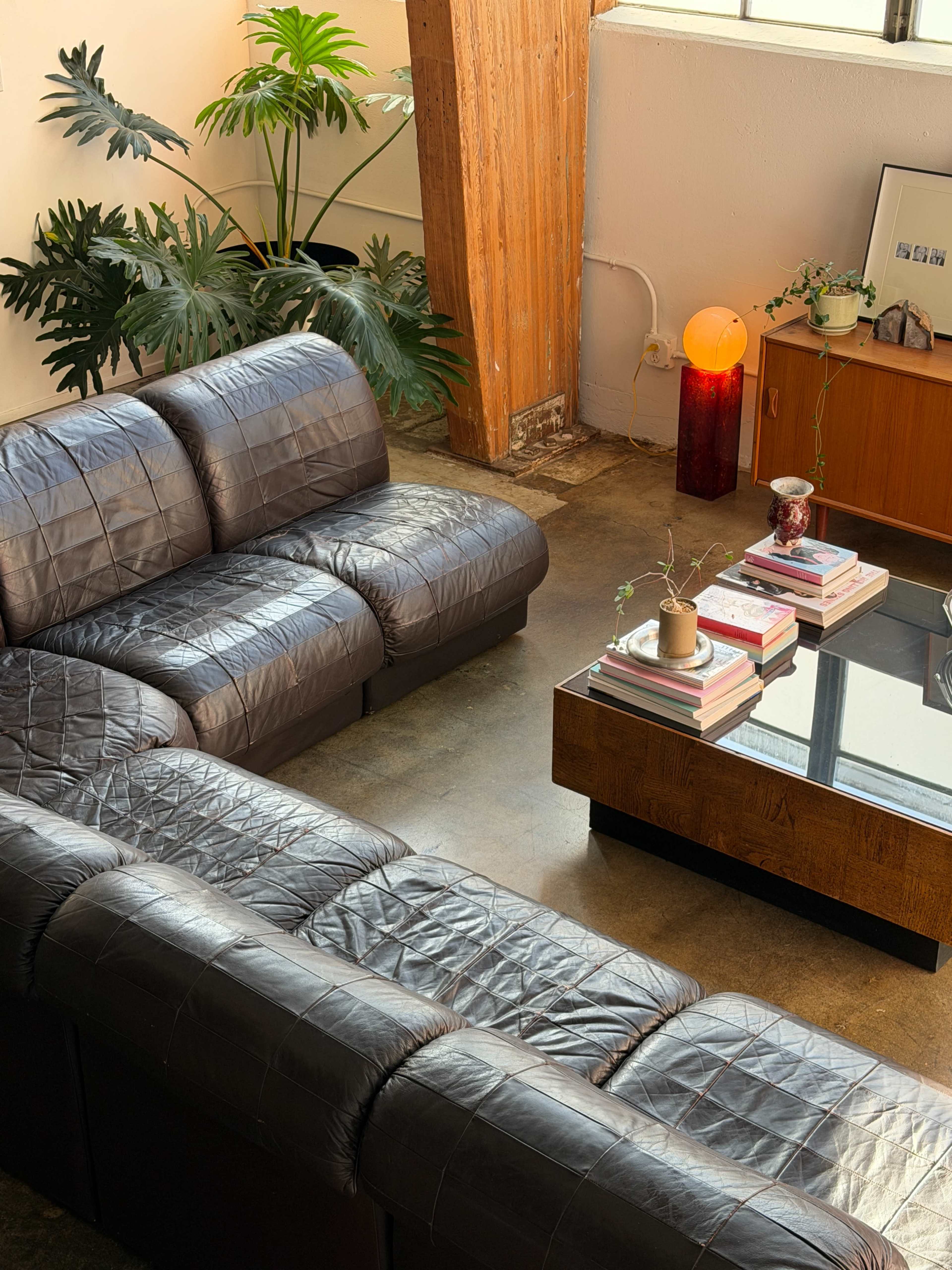 The image shows a modern living room with a brown leather sectional sofa, a glass coffee table stacked with books, and a wooden side table with a lamp and decorative items.