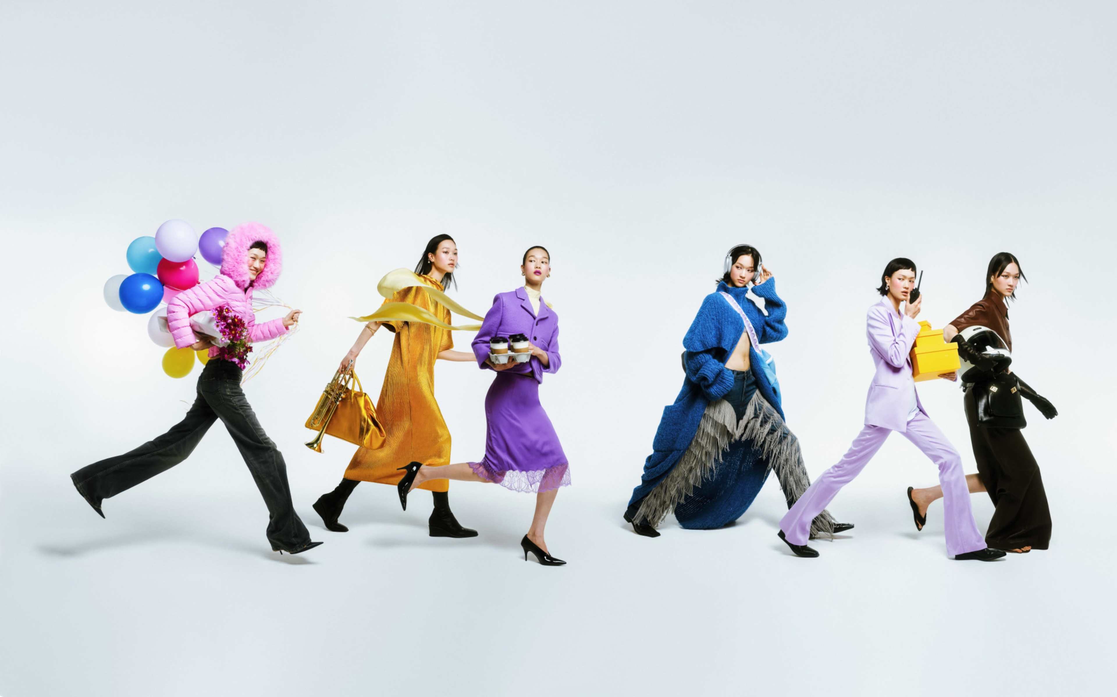 The image features a group of five women, each wearing distinct, colorful outfits and engaged in various activities while walking against a plain white background.