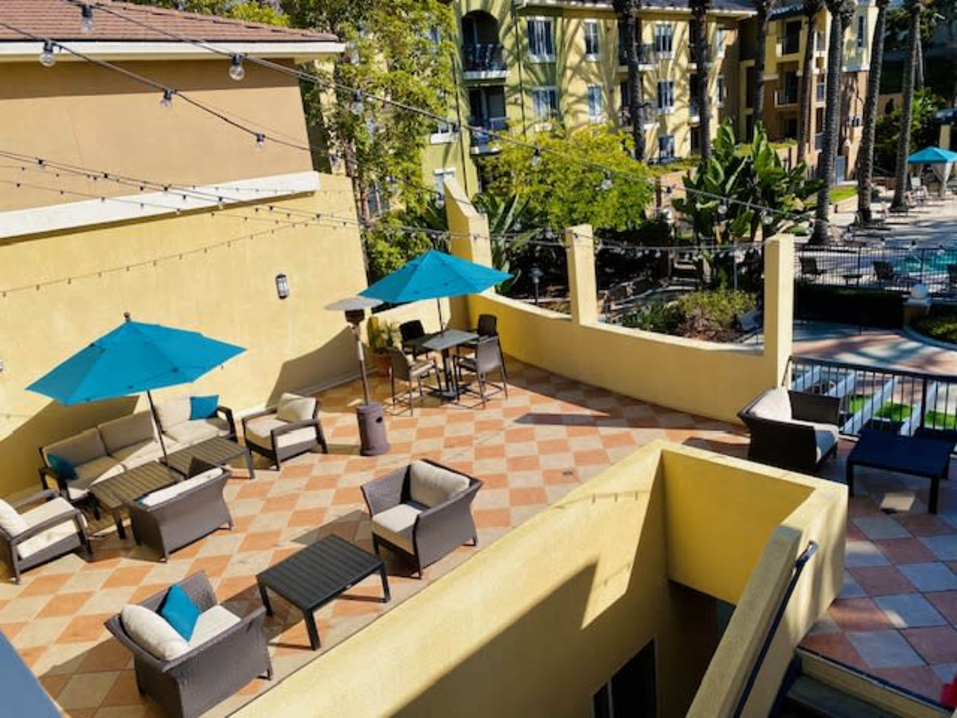 A patio area features several seating arrangements with teal umbrellas and a view of a pool surrounded by greenery.