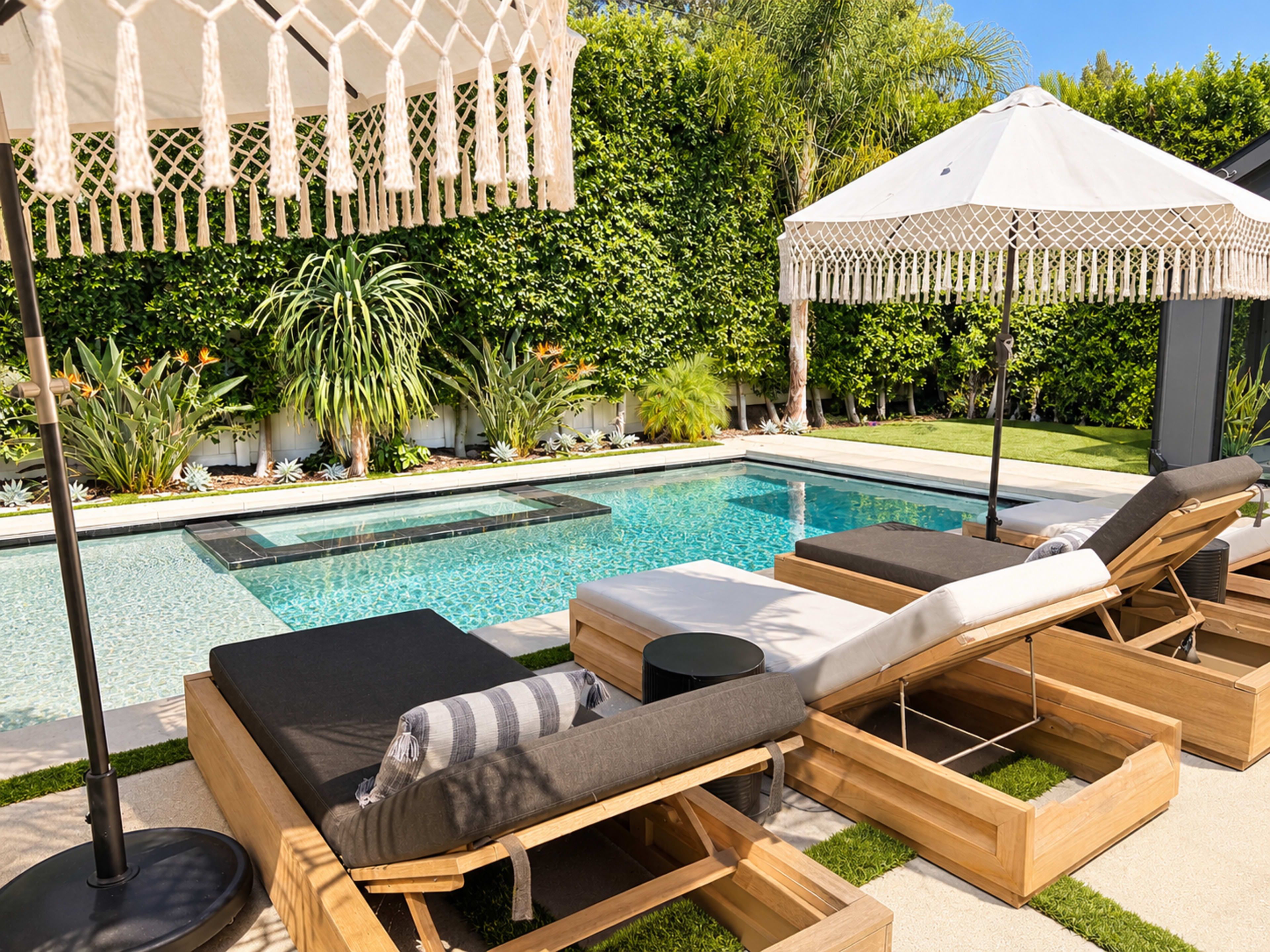 A pair of wooden lounge chairs with cushions are positioned beside a swimming pool surrounded by greenery and patio umbrellas.