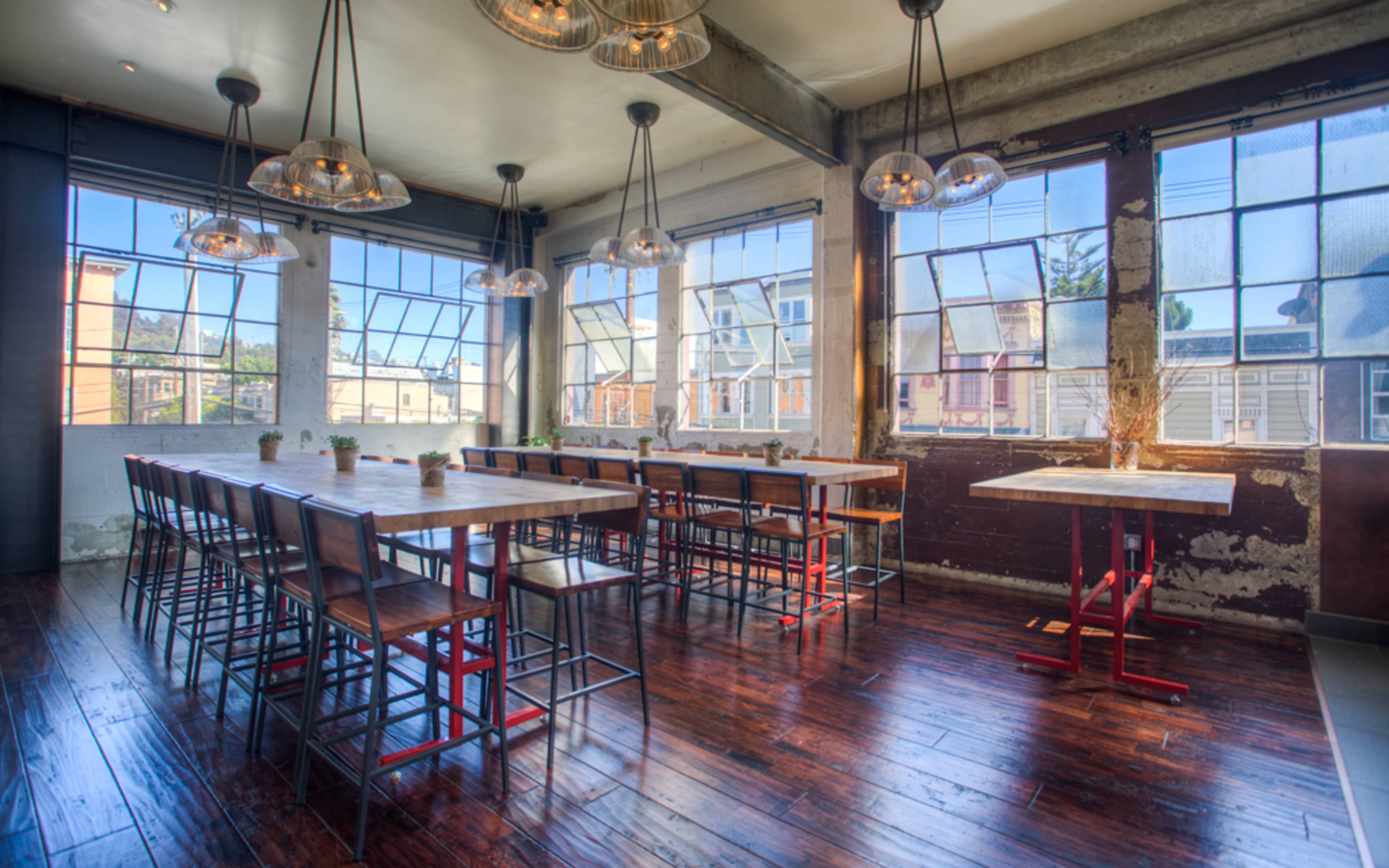 The image shows a spacious dining area with large windows, wooden floors, and long communal tables surrounded by high stools.