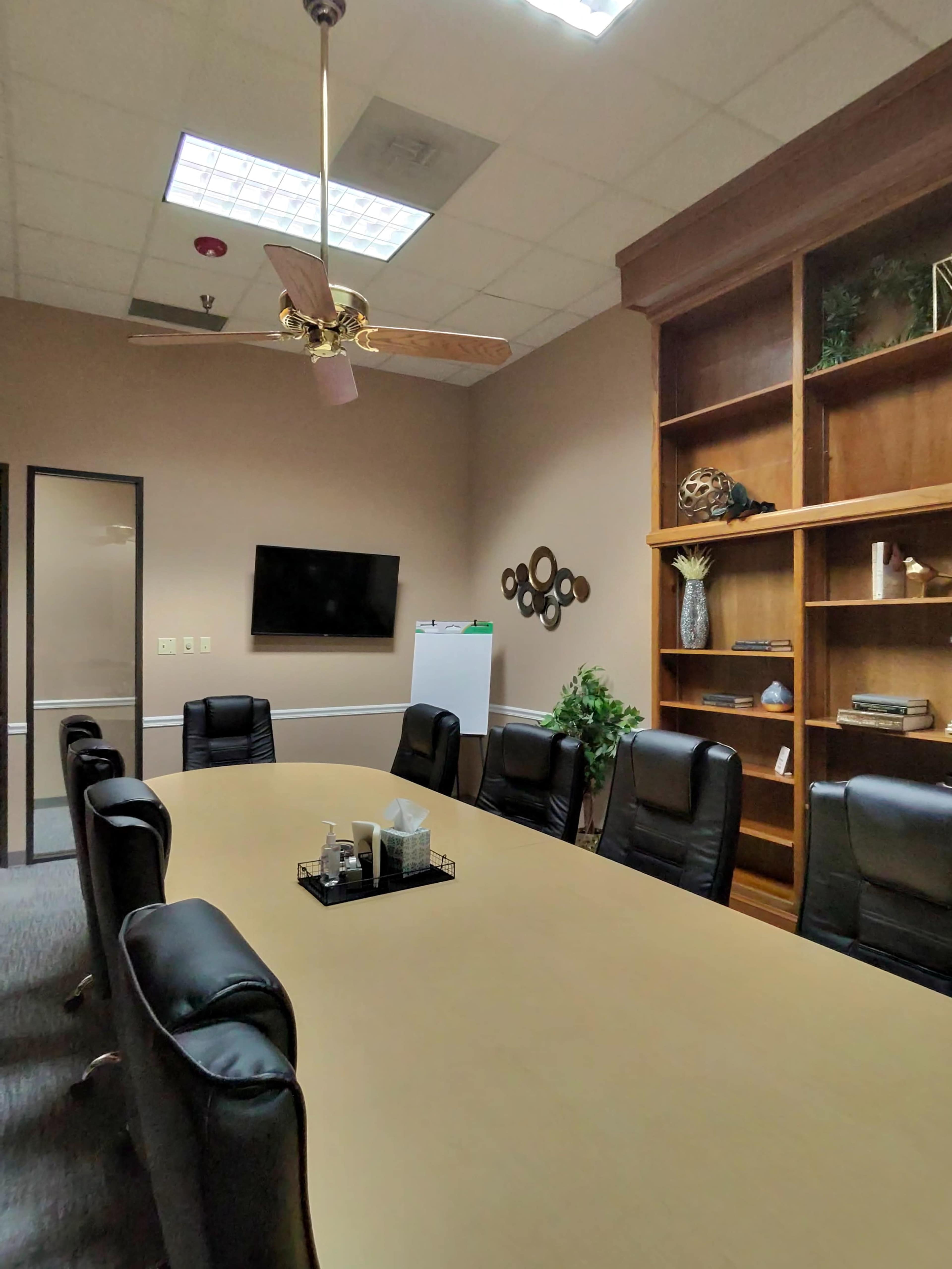 A large conference room features a rectangular table surrounded by black leather chairs, a mounted television, and a bookshelf with decorative items.