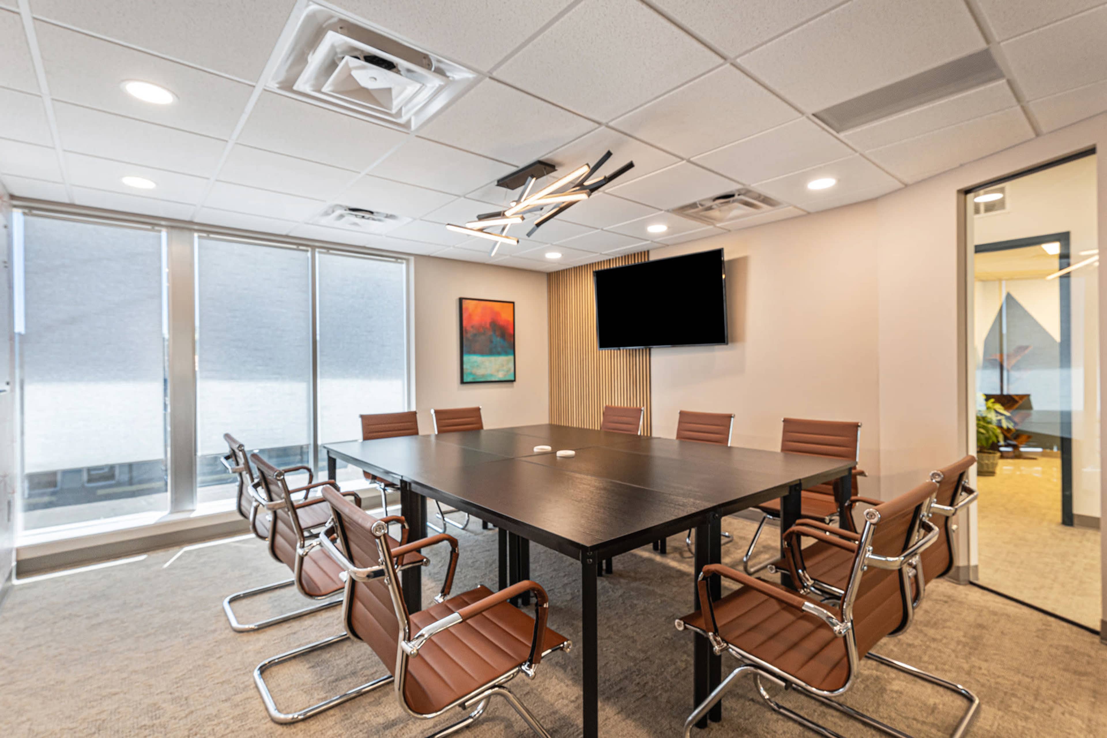 A modern conference room features a large rectangular table surrounded by ergonomic chairs, with floor-to-ceiling windows and a wall-mounted television.