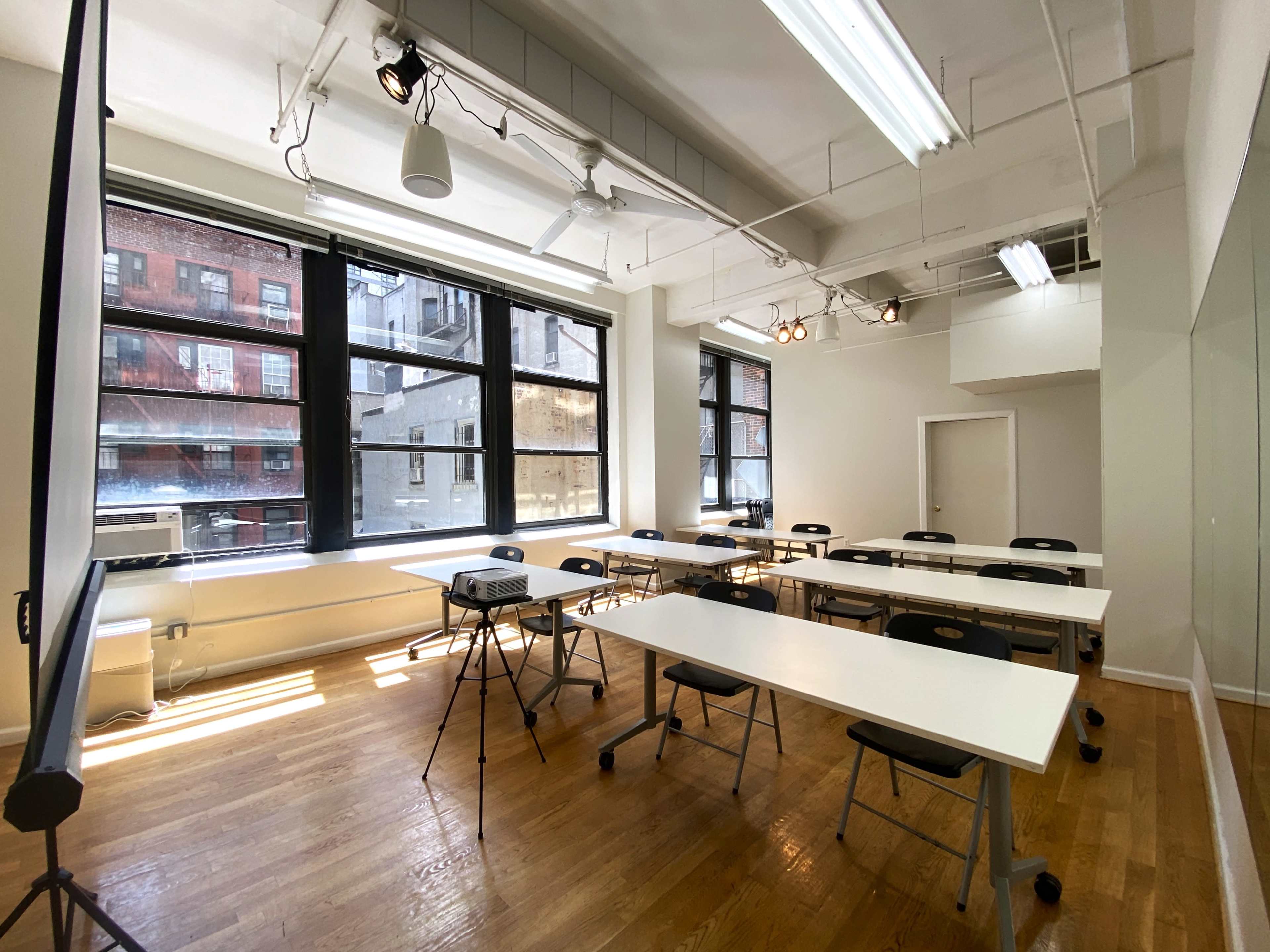 The image shows a brightly lit classroom with white tables and black chairs arranged in rows next to large windows.