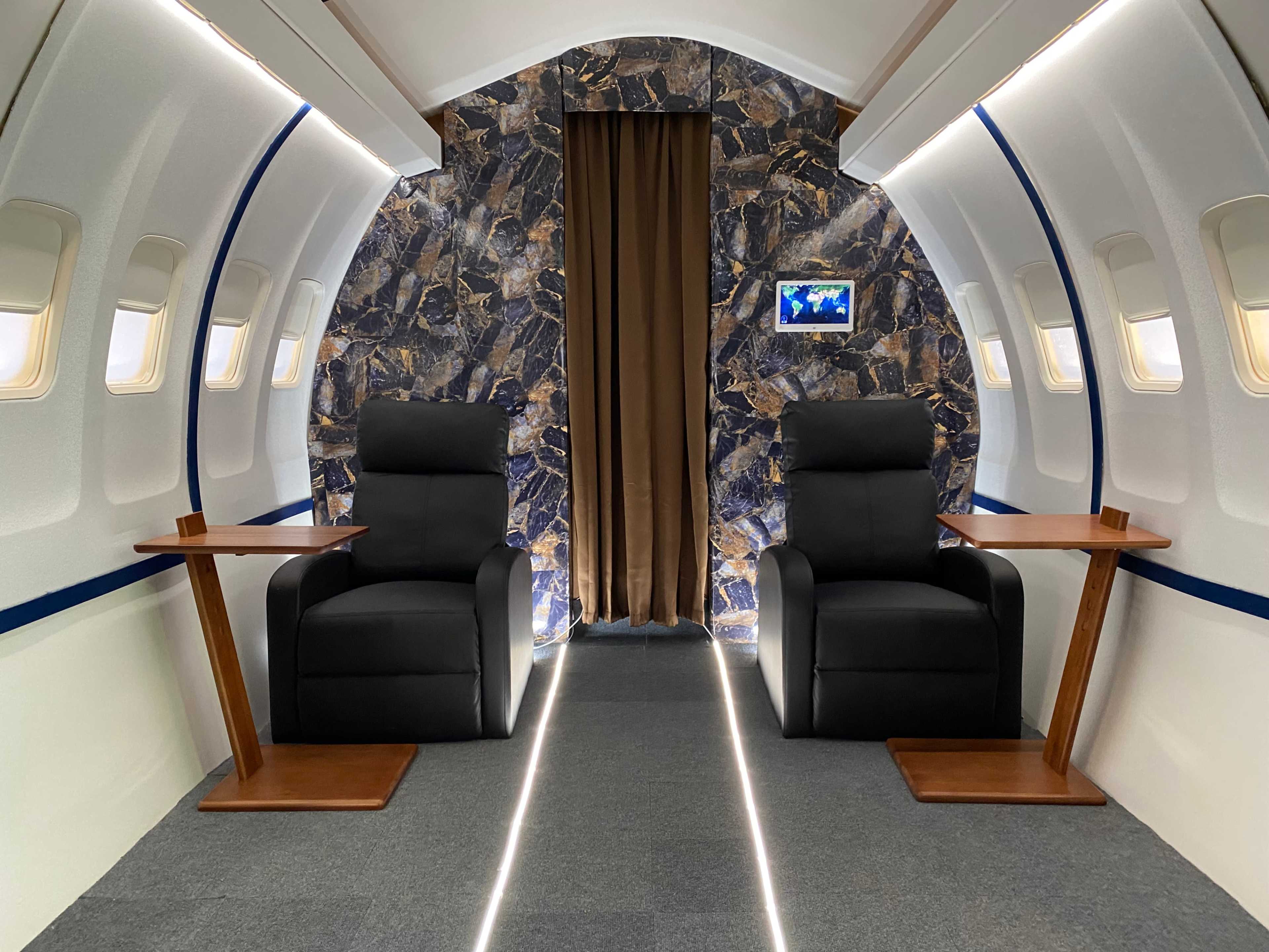 The interior of a luxury airplane cabin featuring two black chairs with wooden side tables, flanked by a decorative stone wall and a curtain.