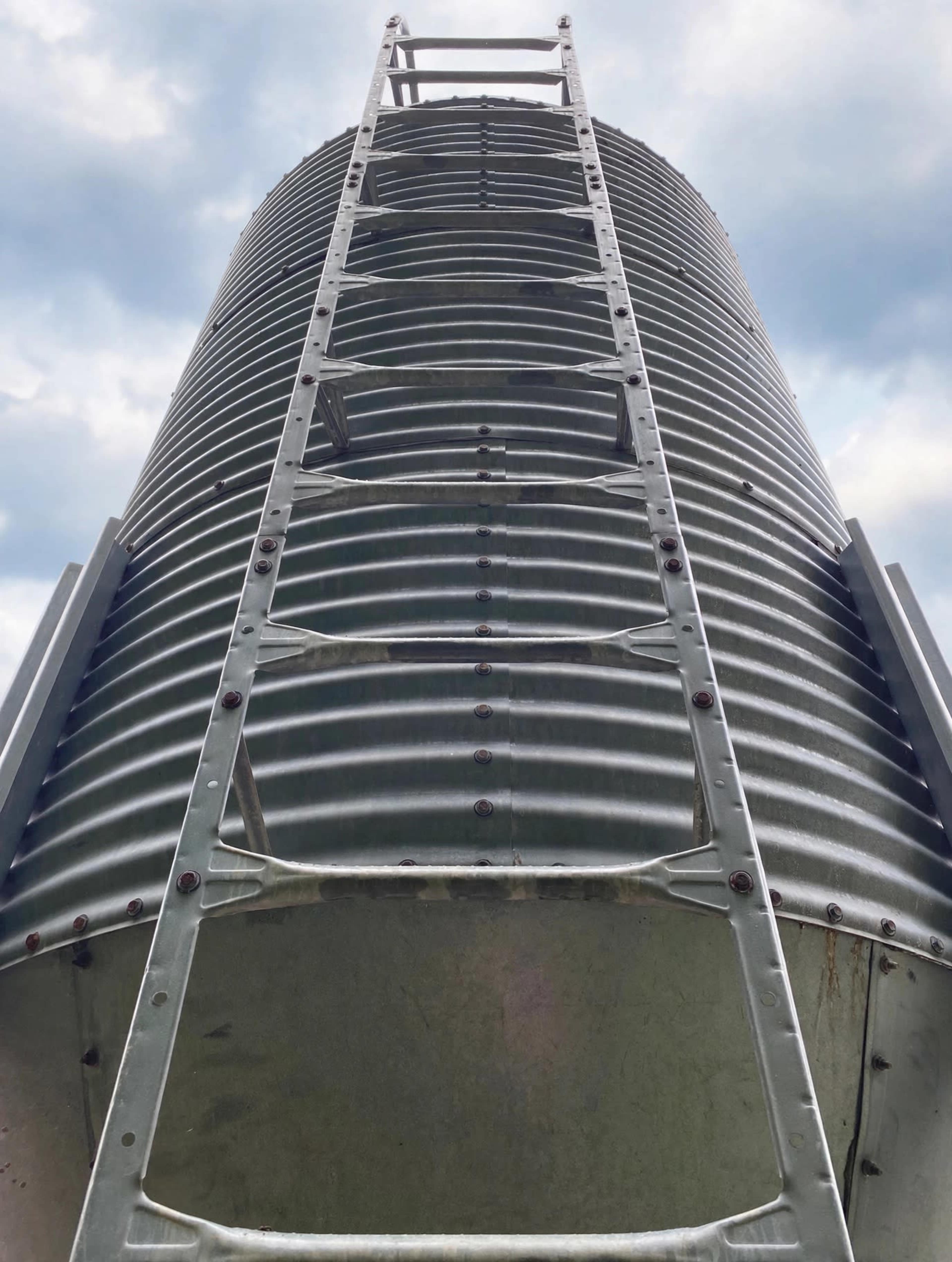 The image shows a tall, cylindrical metal structure with a ladder extending vertically along its side against a cloudy sky.