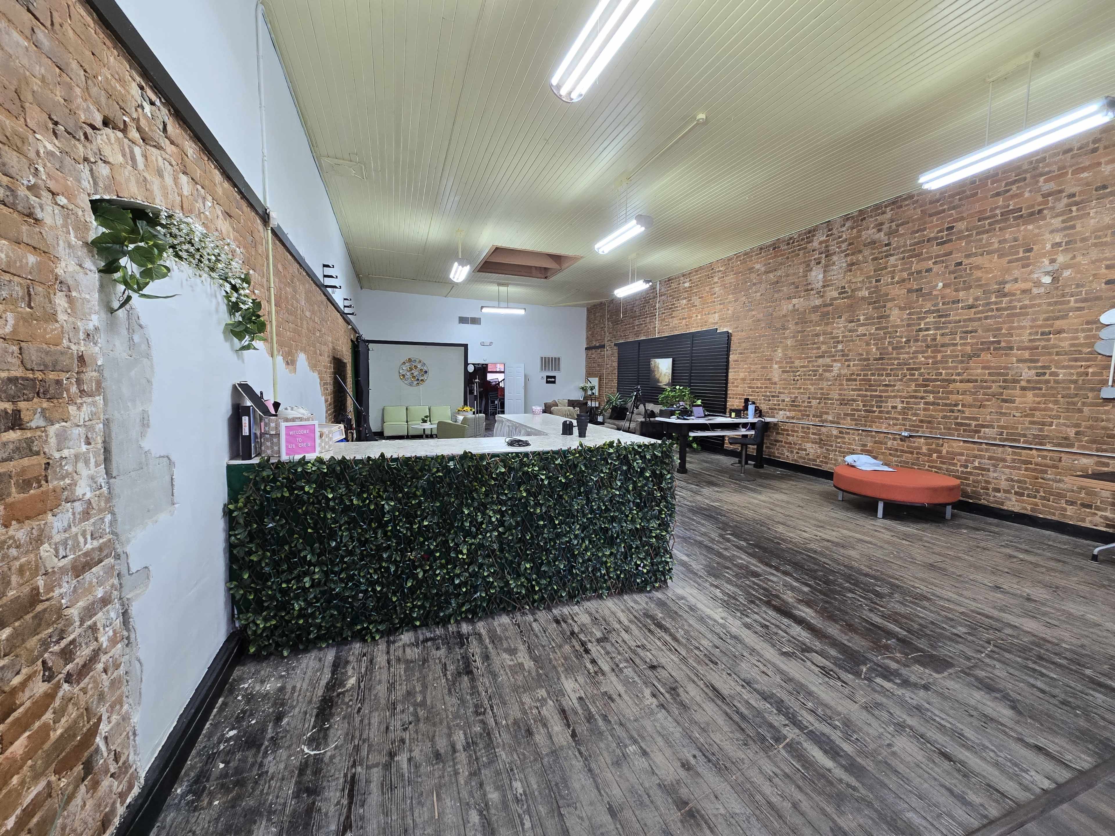 The image shows an interior space with exposed brick walls, wooden floors, and a reception area featuring greenery, a countertop, and a seating area with a round red couch.