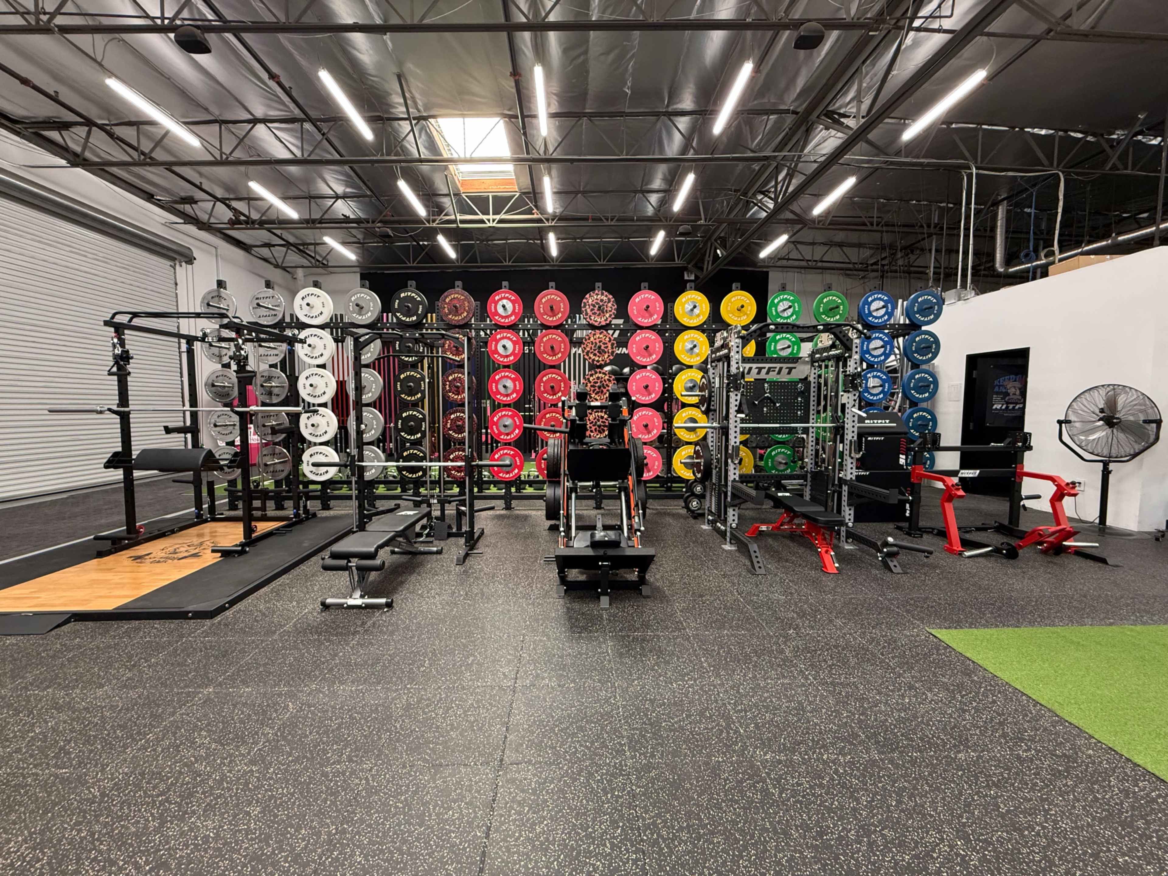 The image shows a well-equipped gym interior featuring various weightlifting machines, racks, and a colorful arrangement of weight plates on a wall.