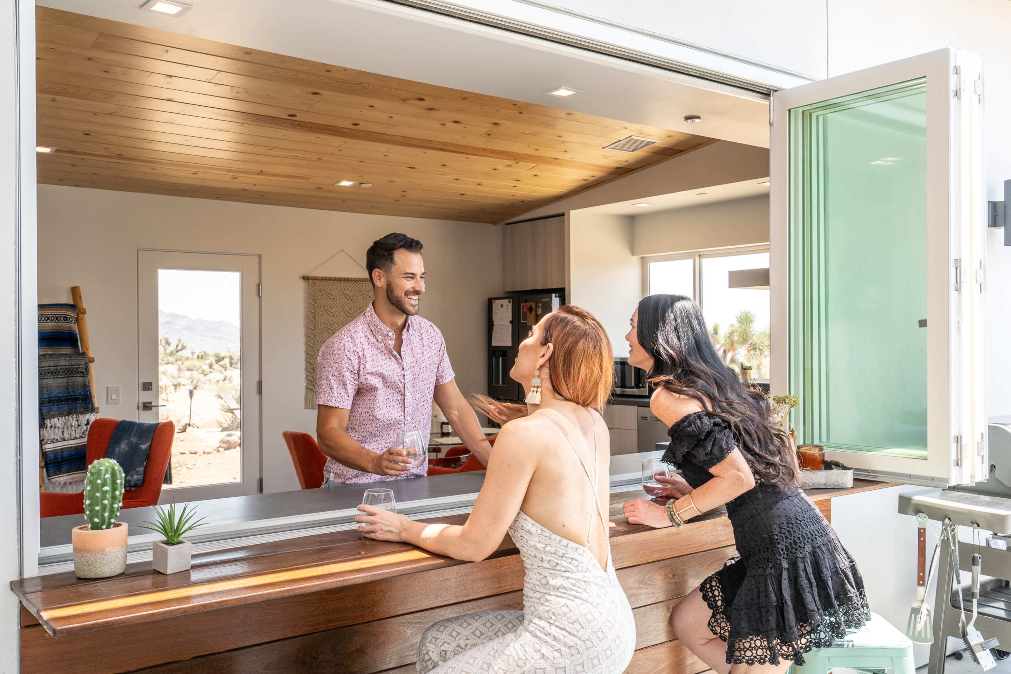 Two women are seated at a kitchen bar, engaging in conversation with a man standing on the opposite side.