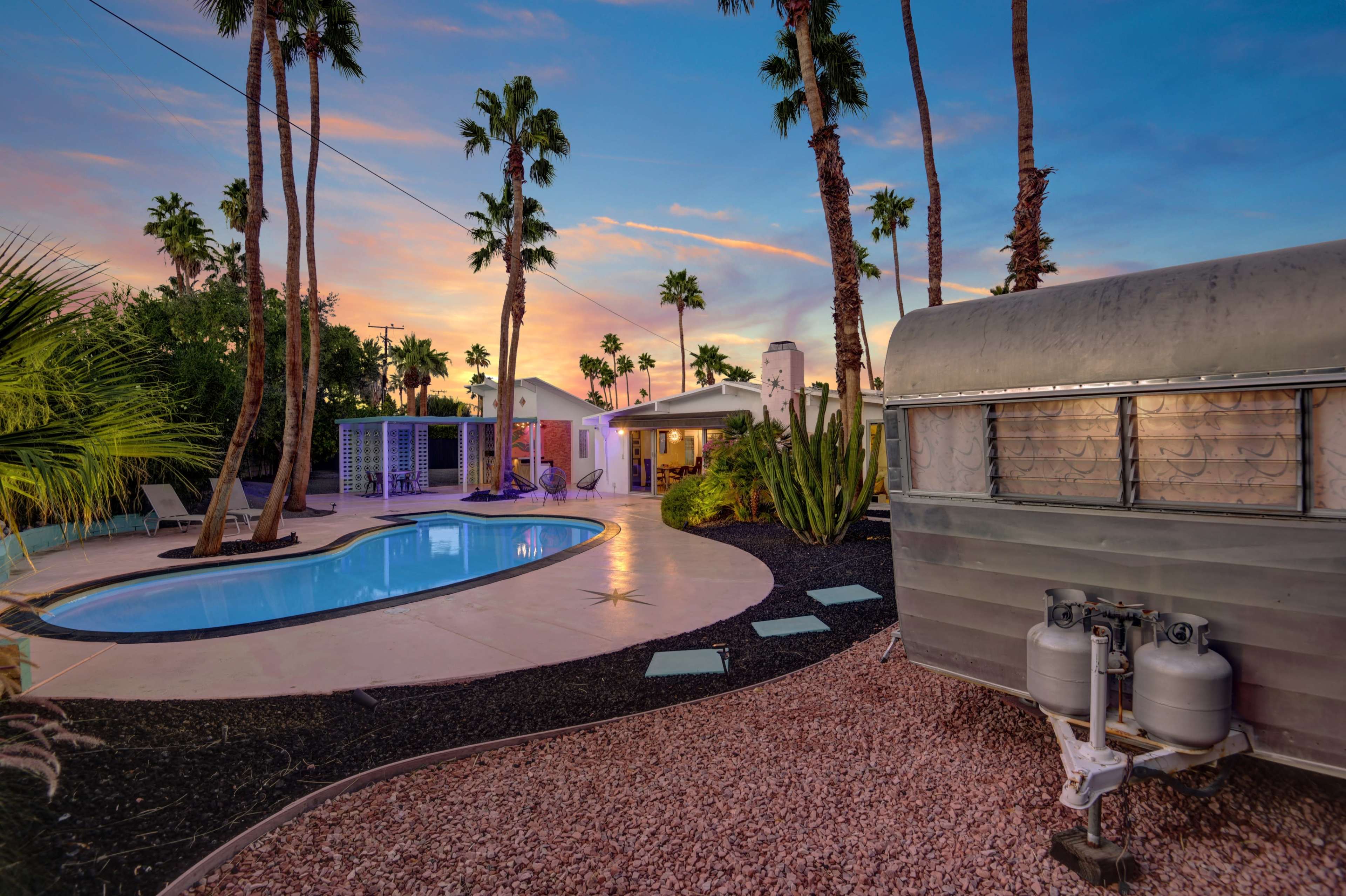 The image features a swimming pool surrounded by a desert landscape, with palm trees and a vintage trailer in the foreground against a colorful sunset.