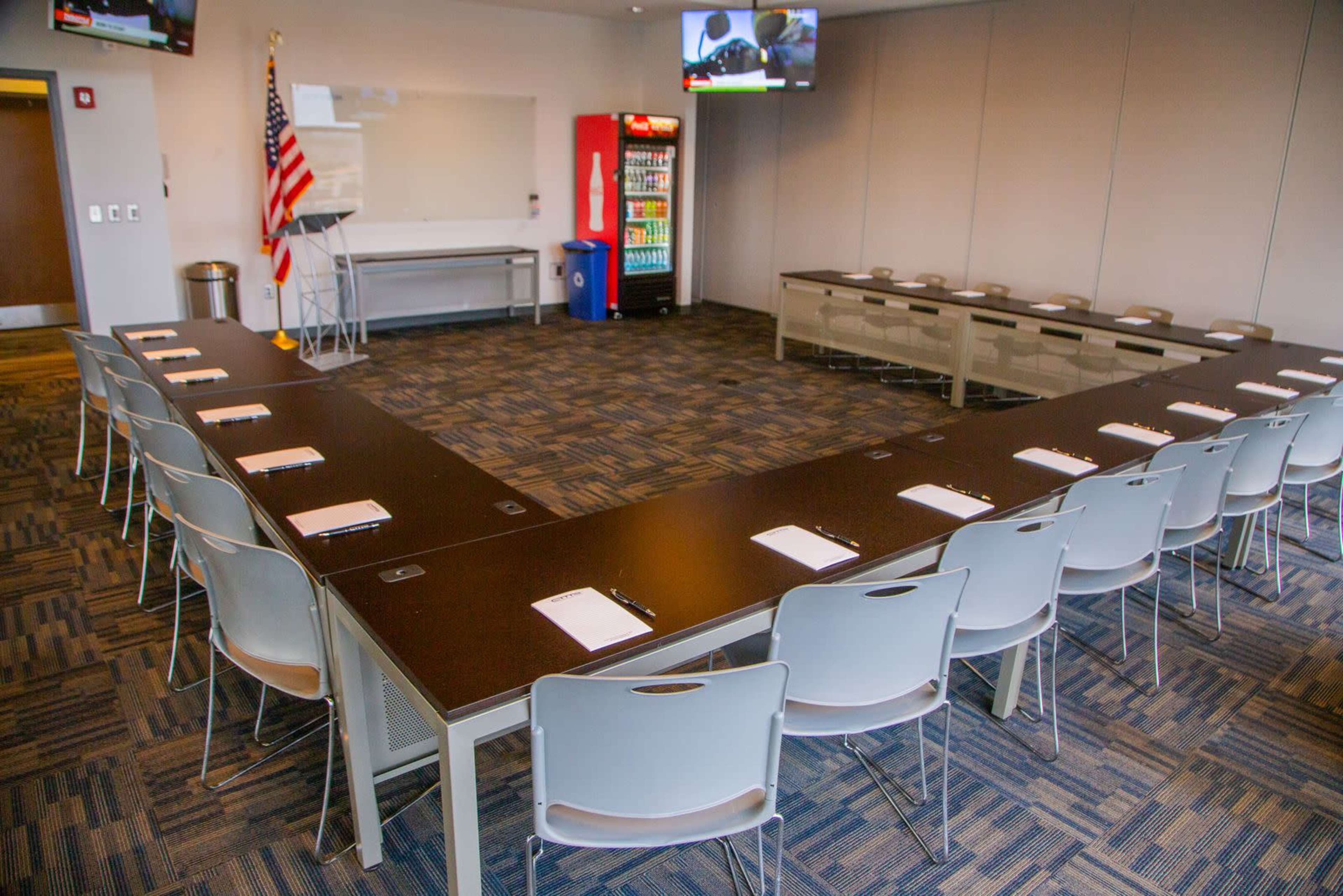 The image shows a meeting room arranged in a U-shape with a large table, chairs, and a vending machine in the corner.