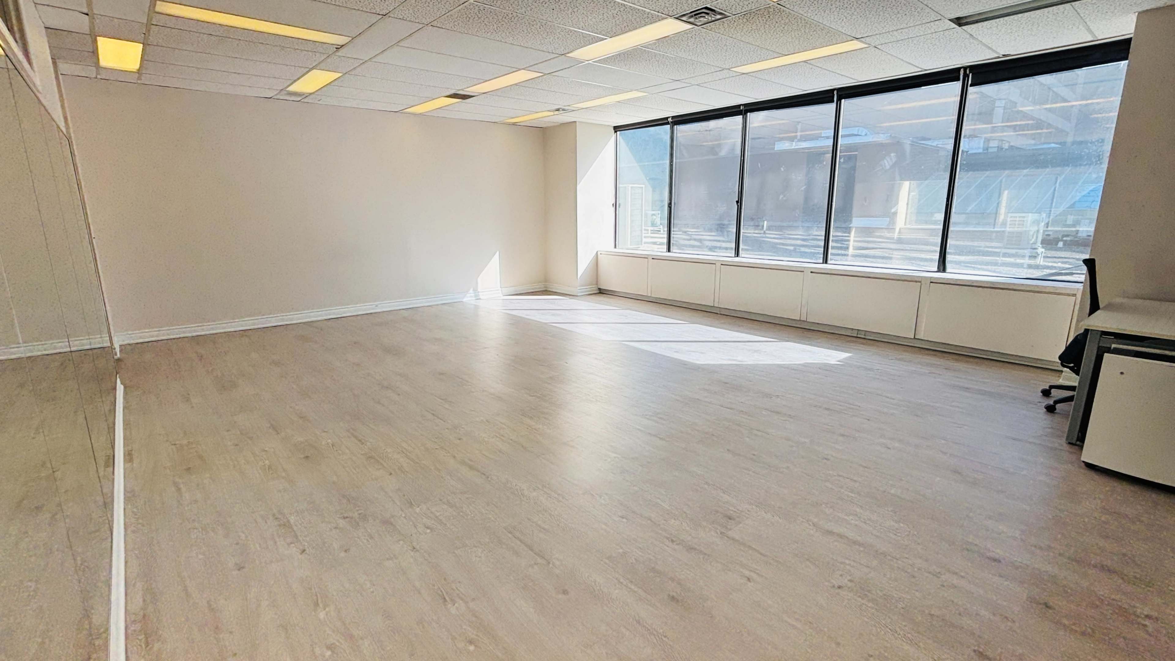 The image shows a spacious, empty room with large windows allowing natural light to fill the space, and a polished wooden floor.