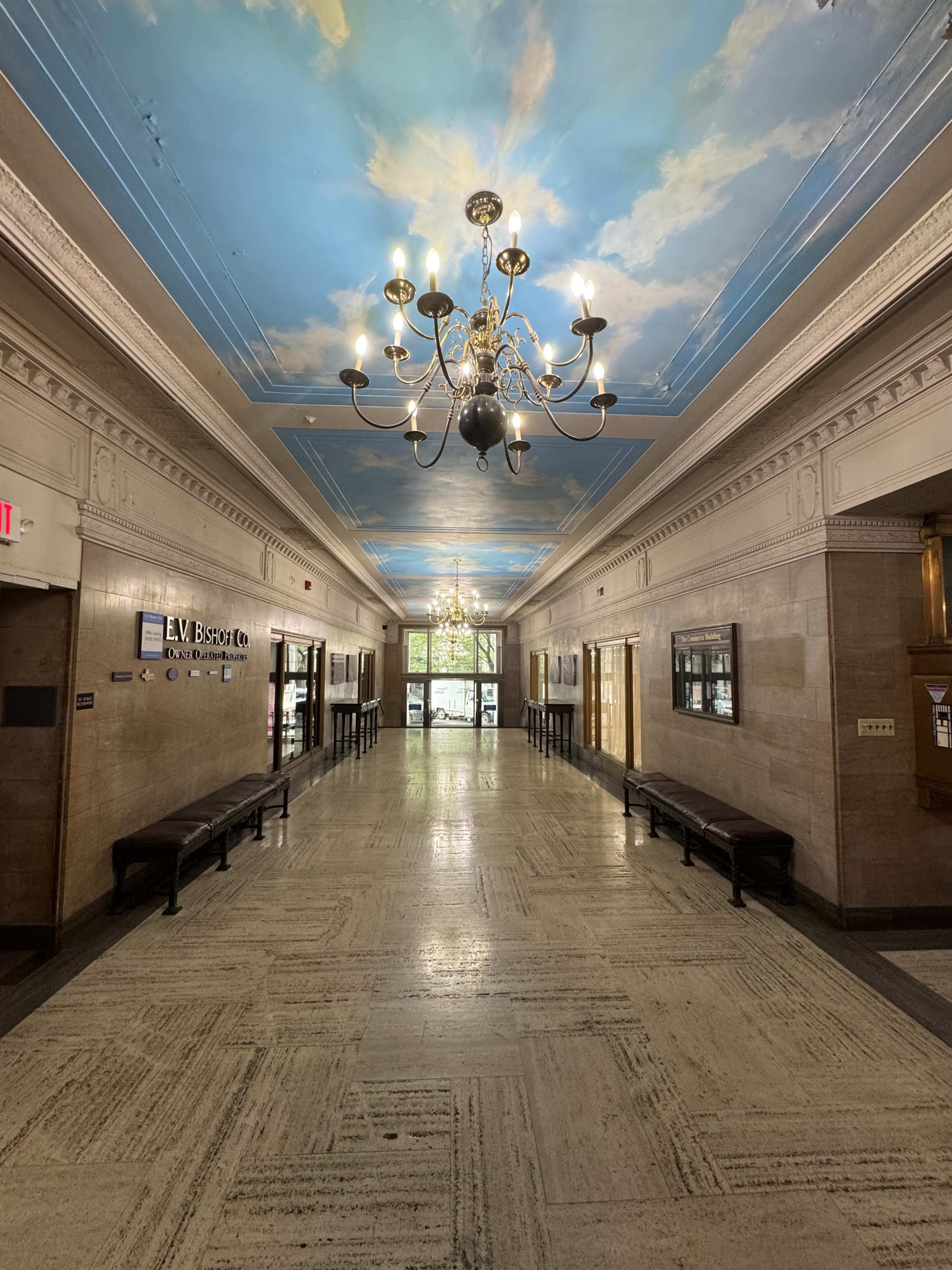 A long hallway featuring a painted ceiling with clouds, illuminated by chandeliers, and lined with benches on either side.