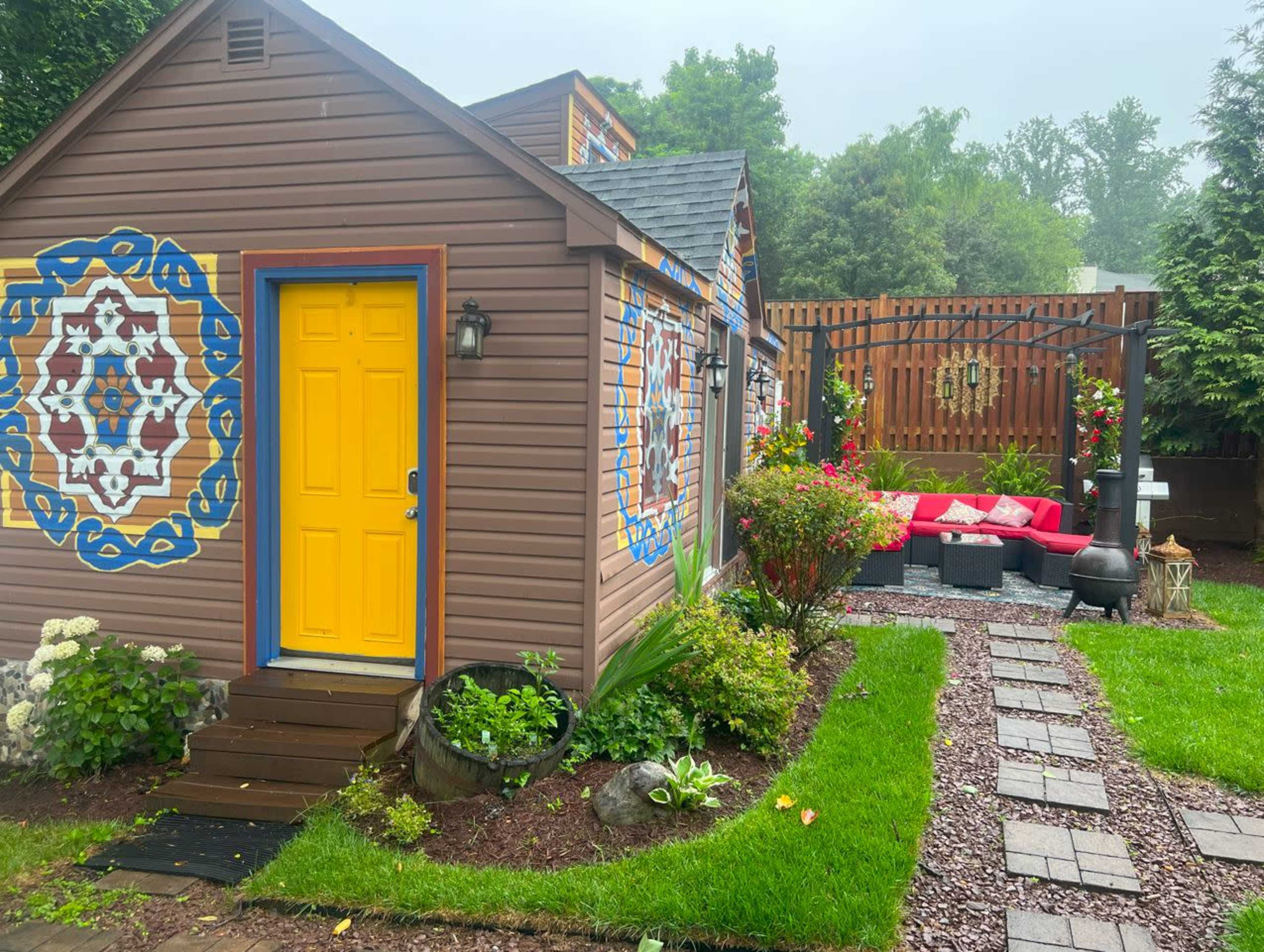 A small wooden house with colorful designs on the exterior features a bright yellow door and a landscaped garden with a stone pathway leading to a seating area.