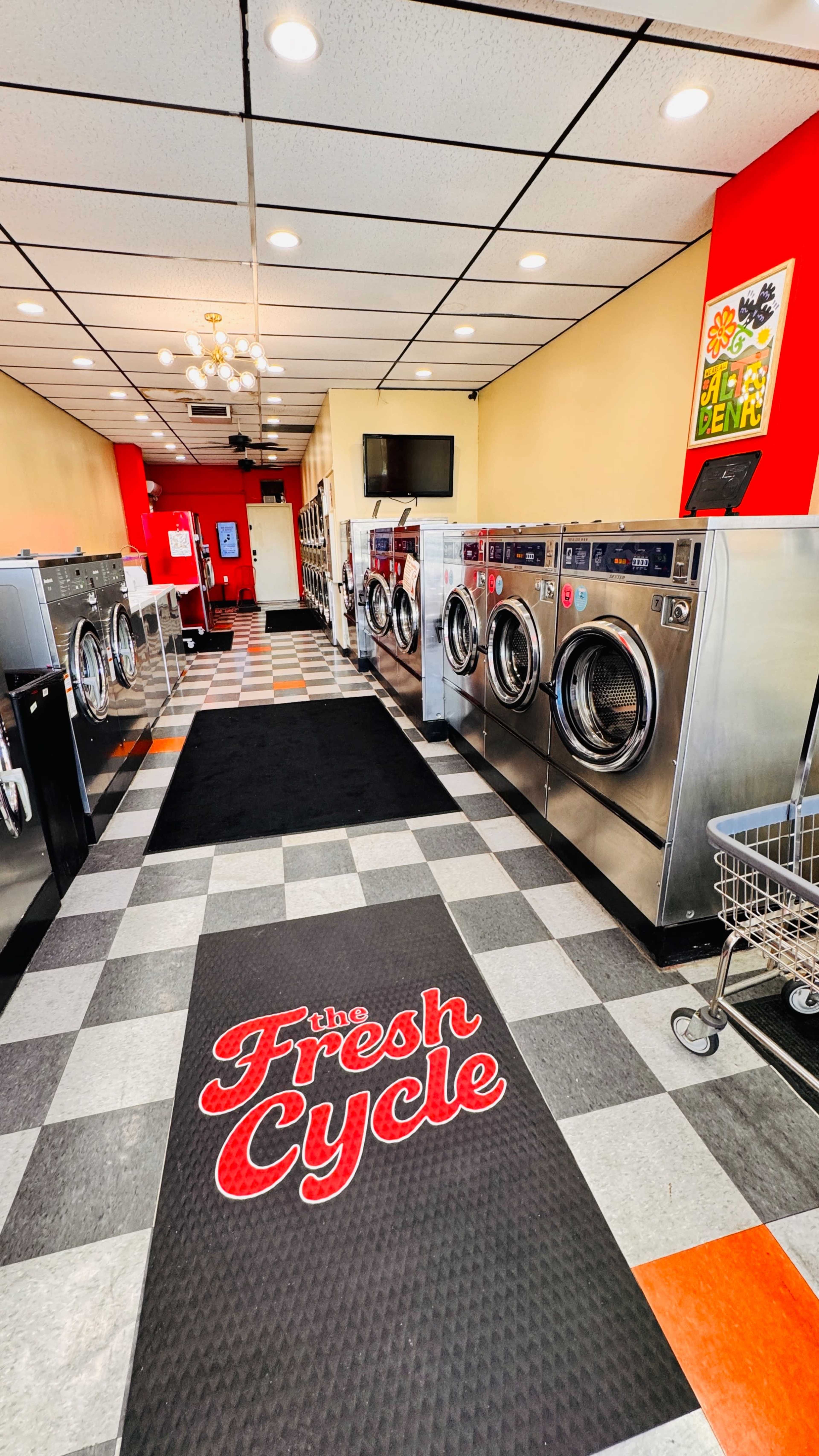 The image shows the interior of a laundromat called "the Fresh Cycle," featuring rows of washing machines and a black carpet at the entrance.