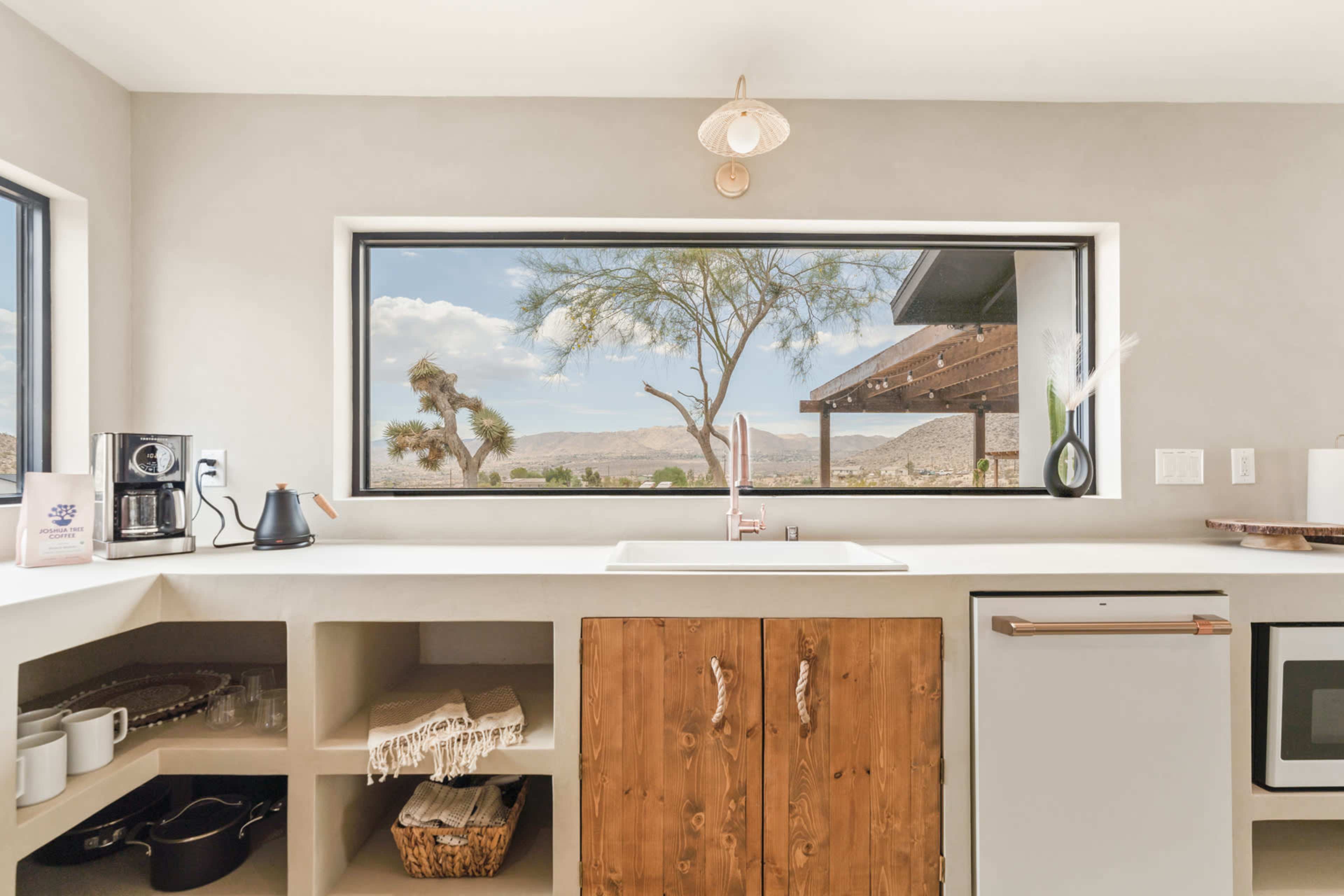 A modern kitchen with a large window overlooking a desert landscape features a sink and wooden cabinetry.