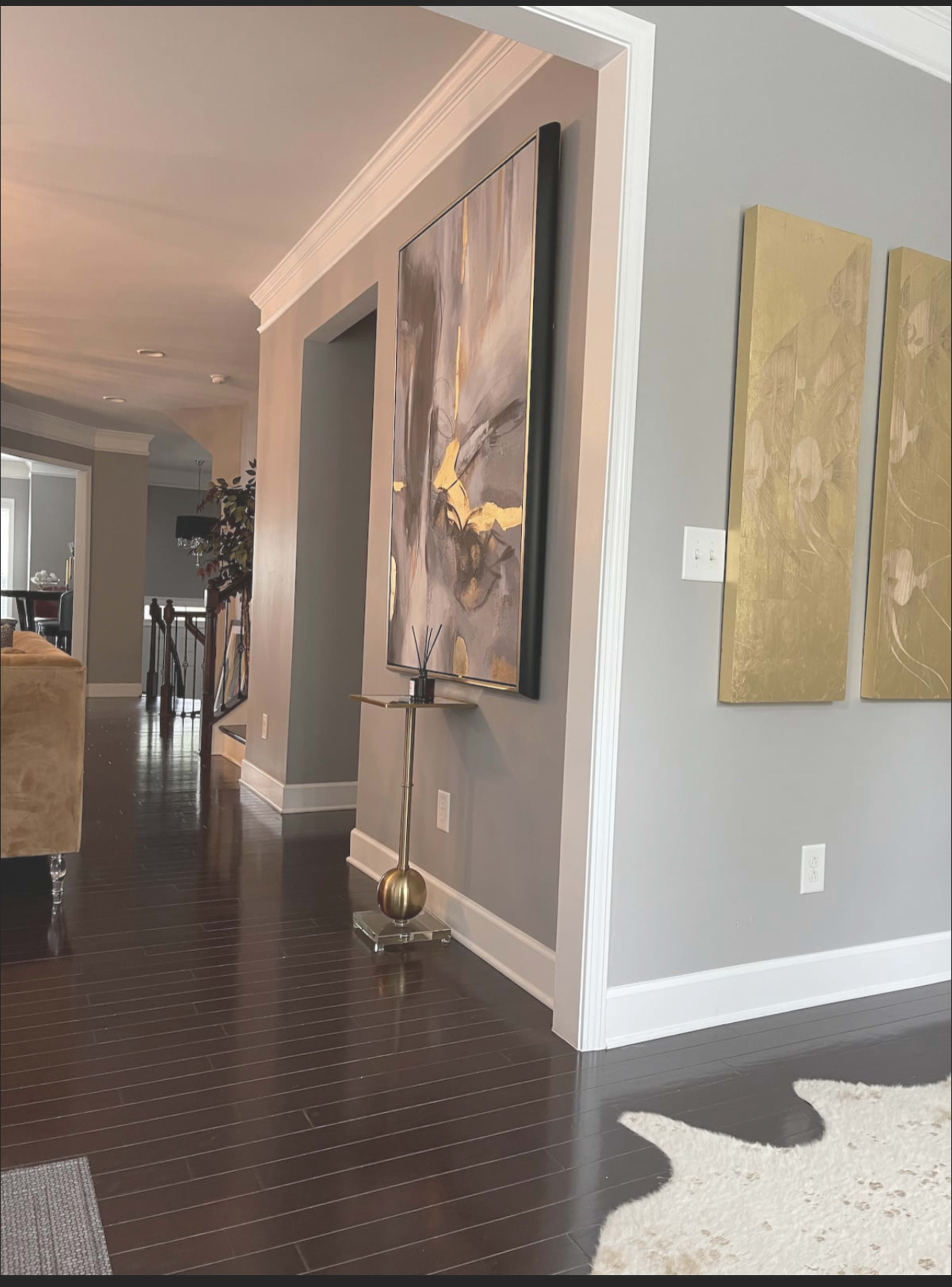 A modern living space with a gray wall, a large abstract painting, and two gold-framed artworks, alongside a dark wooden floor and a small decorative table.
