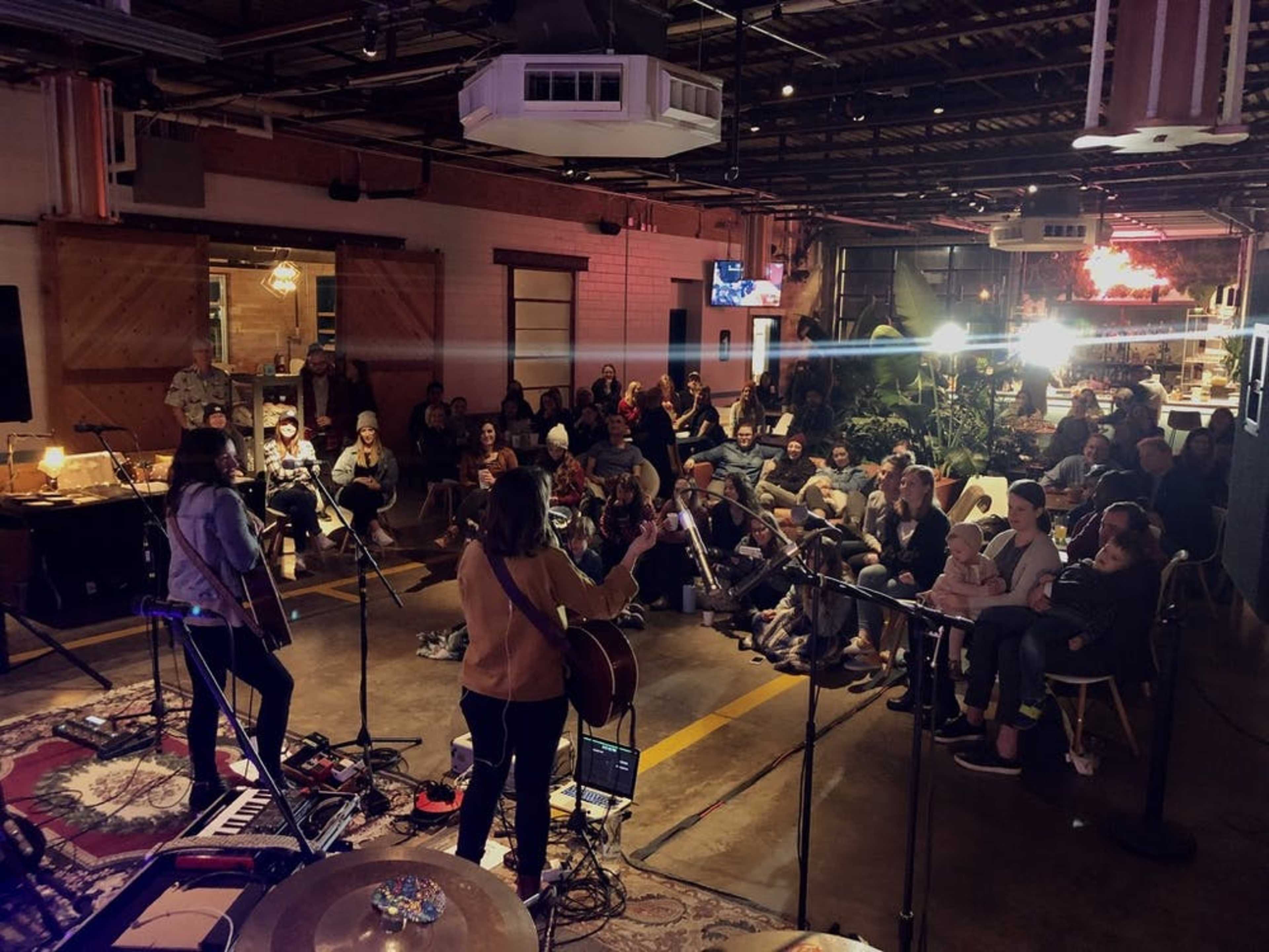 Two musicians perform on stage in a dimly lit venue while an audience sits facing them.