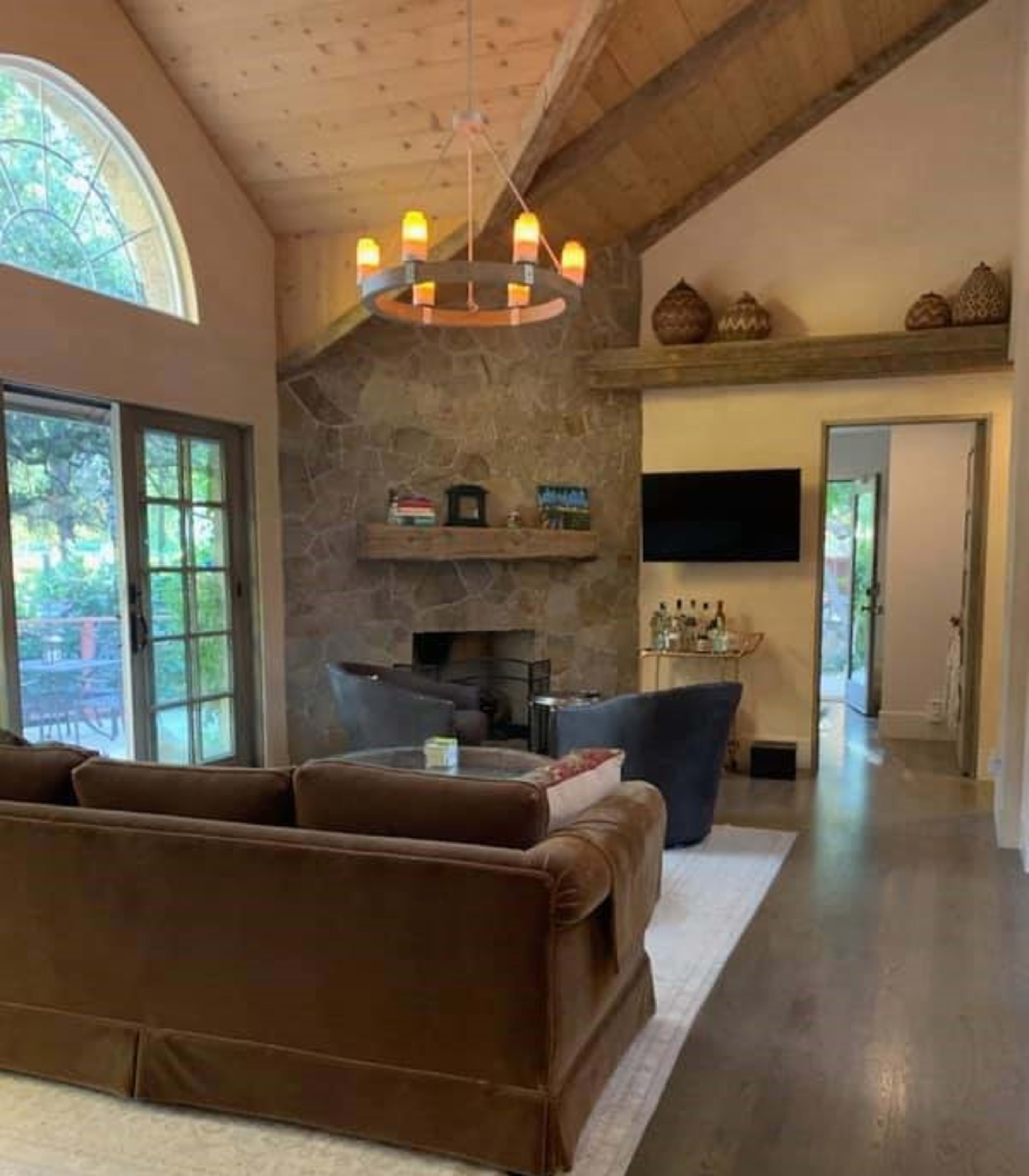 A cozy living room with a stone fireplace, wooden beams, and large windows, featuring a brown sofa and a small bar area.