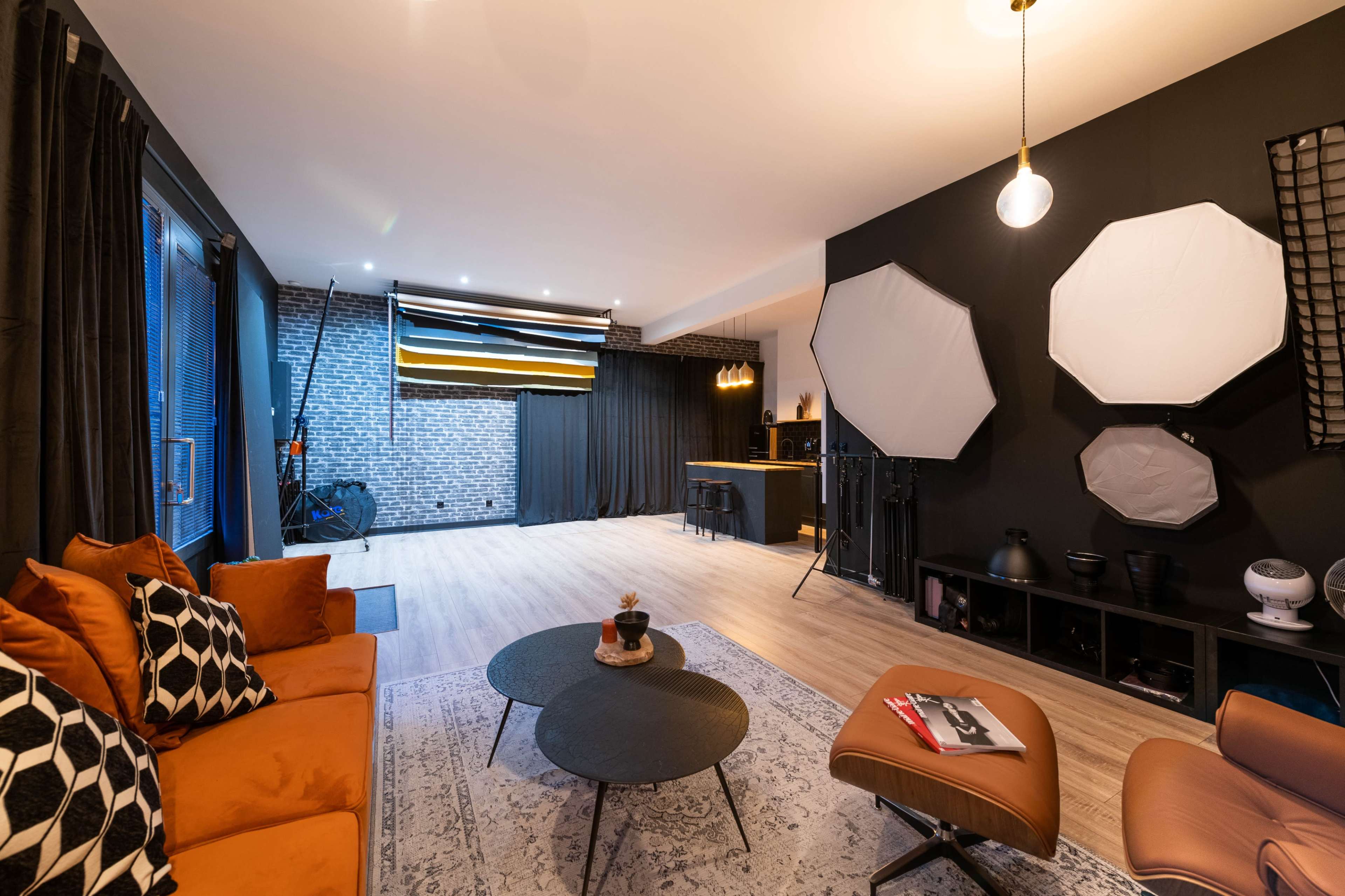 The image shows a modern studio space featuring a black accent wall, large light modifiers, a cozy seating area with orange cushions, and wooden flooring.