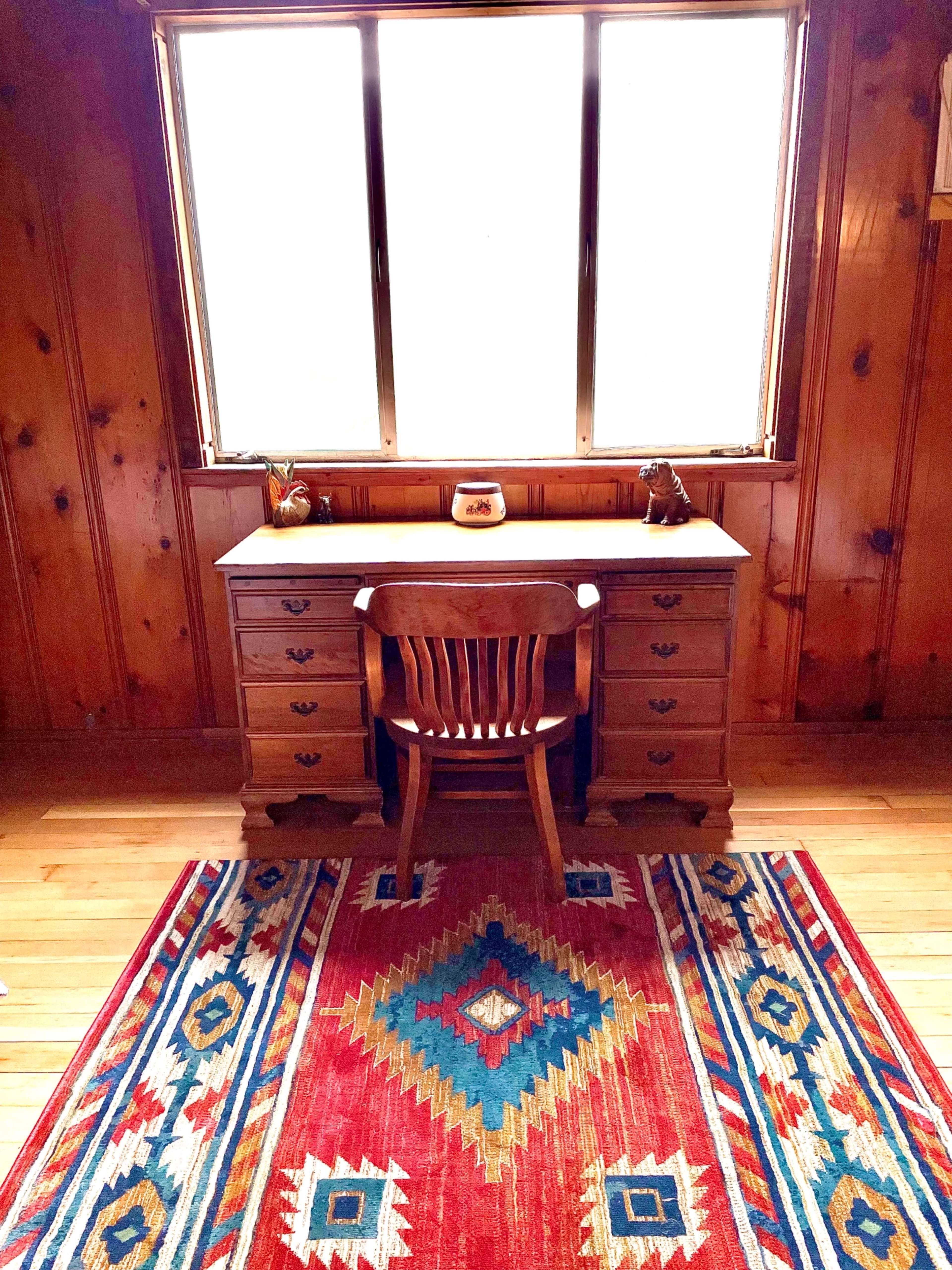A wooden desk with a chair is positioned in front of a large window, on a patterned red and blue rug in a room with wood-paneled walls.